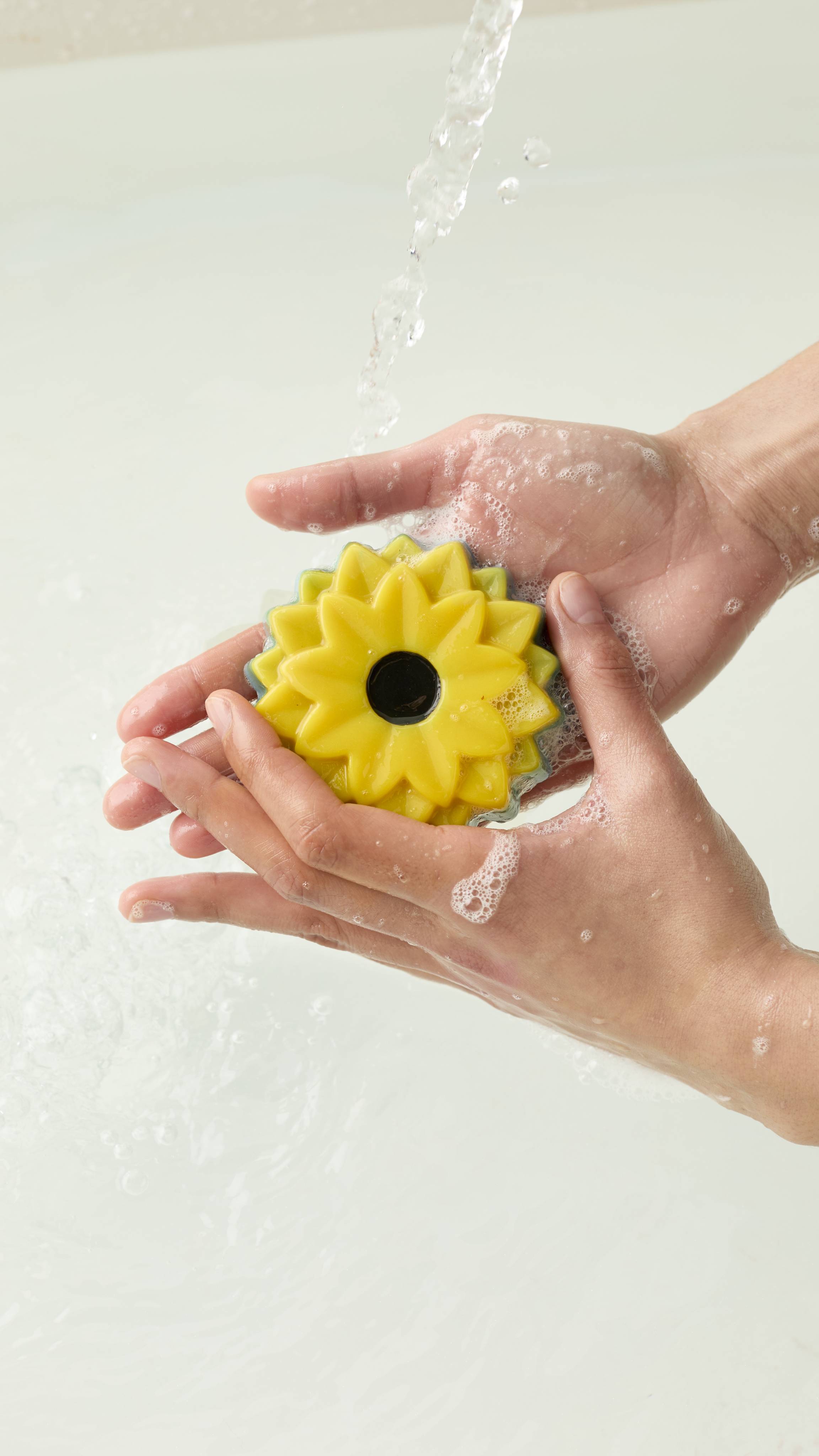 An aerial view of two hands holding the sunflower-shaped There Will Be Sun soap. Water runs over the hands as they create a soapy lather with the product.