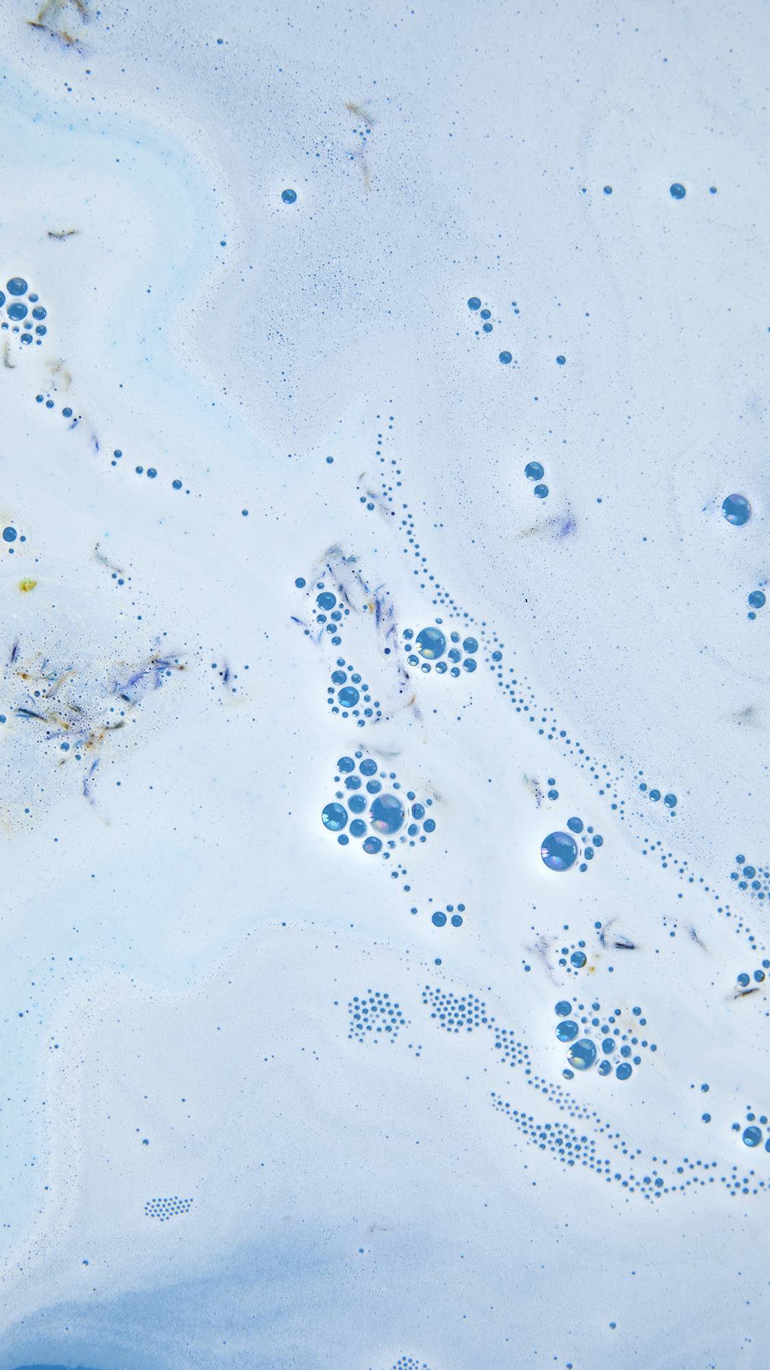 Overhead close-up of light blue bathwater with swirling white foam, small clusters of bubbles, and floating bits of dried blue petals.
