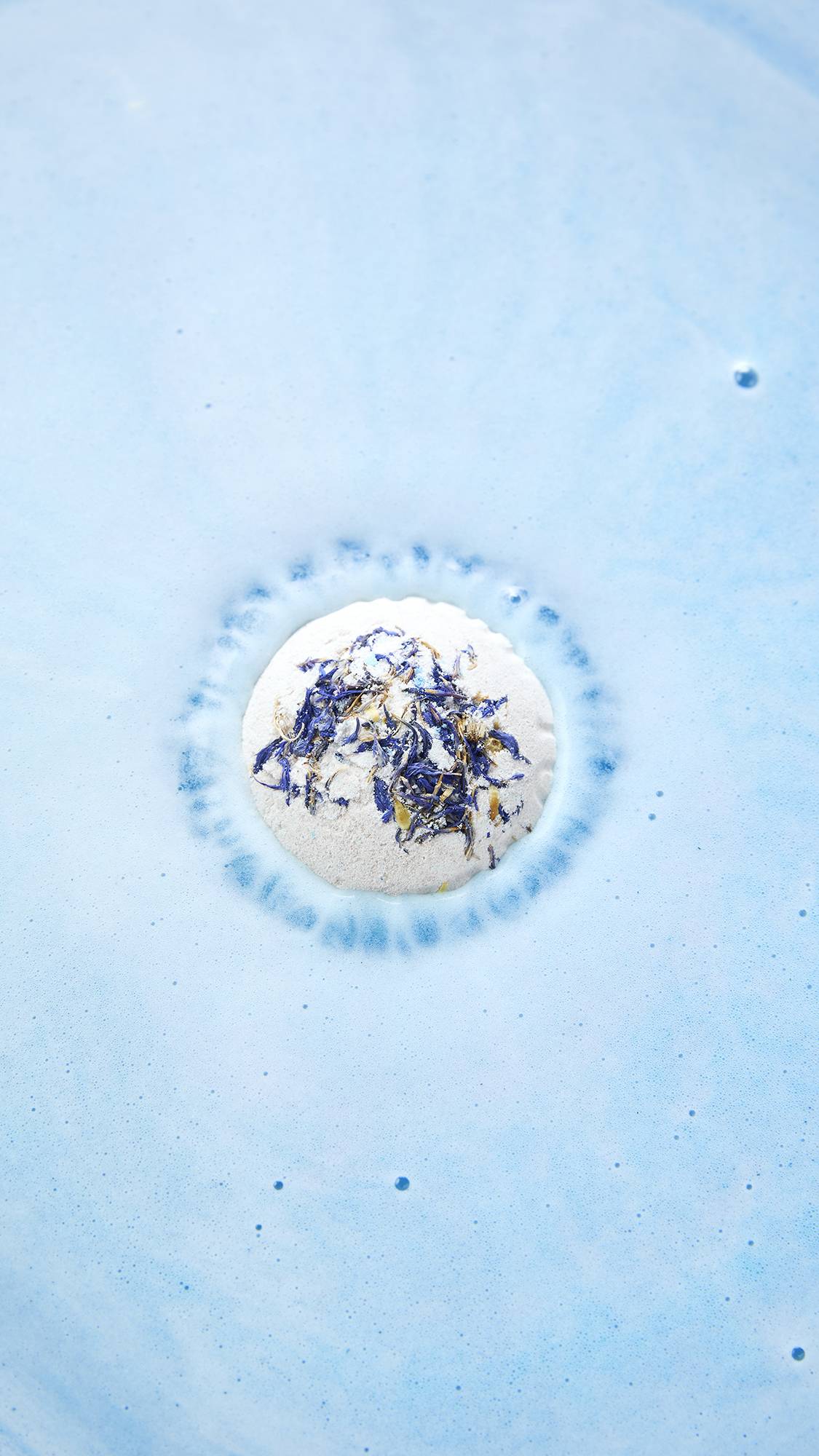 An aerial view of a LUSH "This Too Shall Pass" bath bomb topped with dark blue cornflower petals dissolving in light blue bathwater, creating a ring of blue fizz and bubbles around itself.