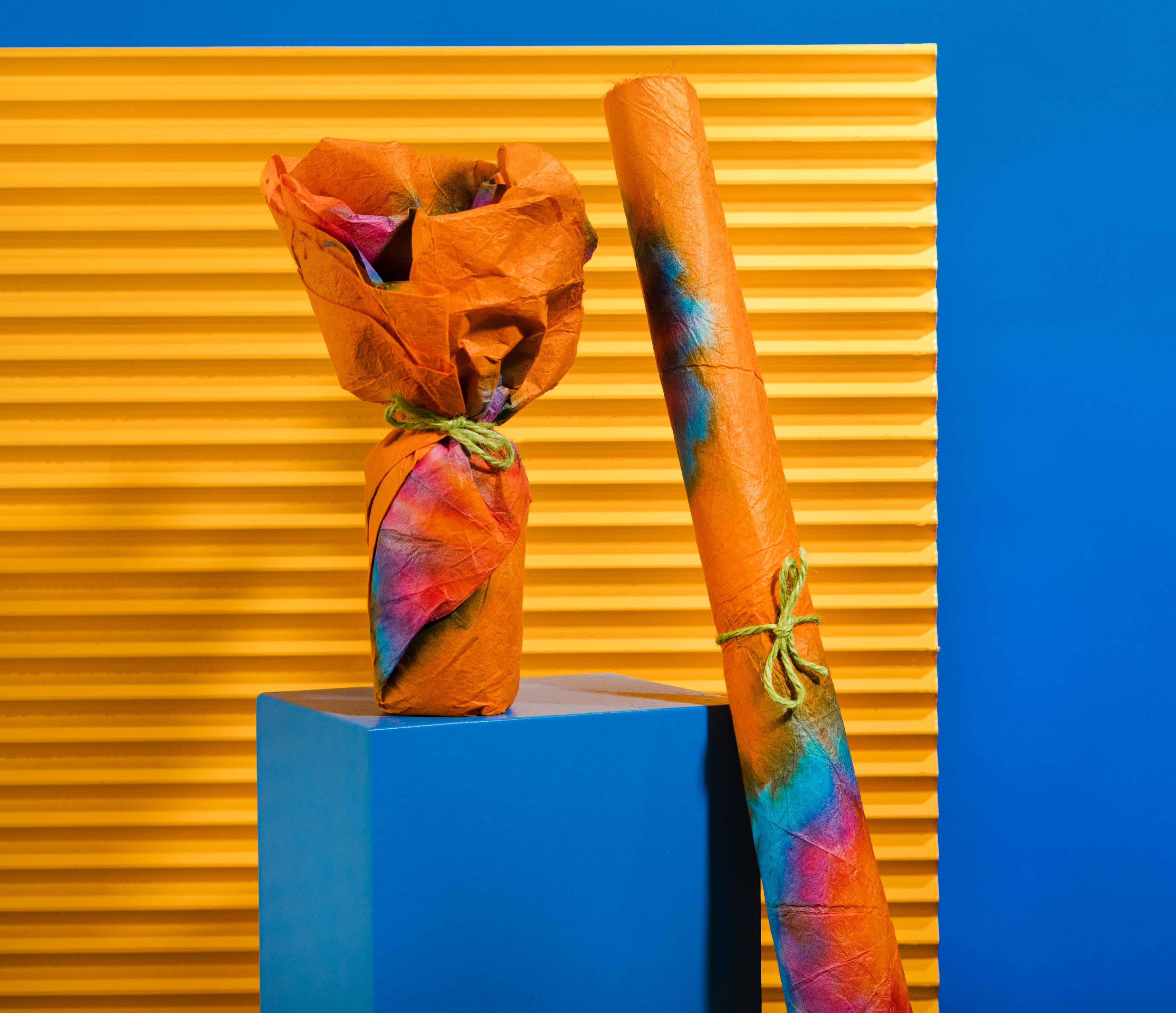 The Lokta paper is wrapped around a product, and rolled up and tied with string, on a blue plinth before an orange background.