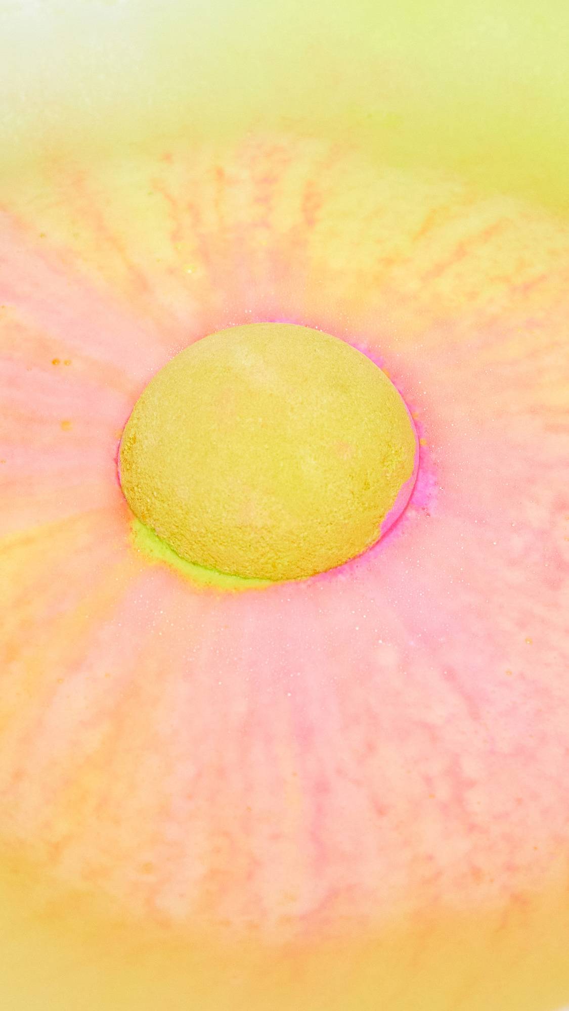 The Time Of The Season bath bomb has been placed into the bath water where it sits like a giant round sun in the centre shooting off swirls of bright yellow and subtle pink like a sunset.