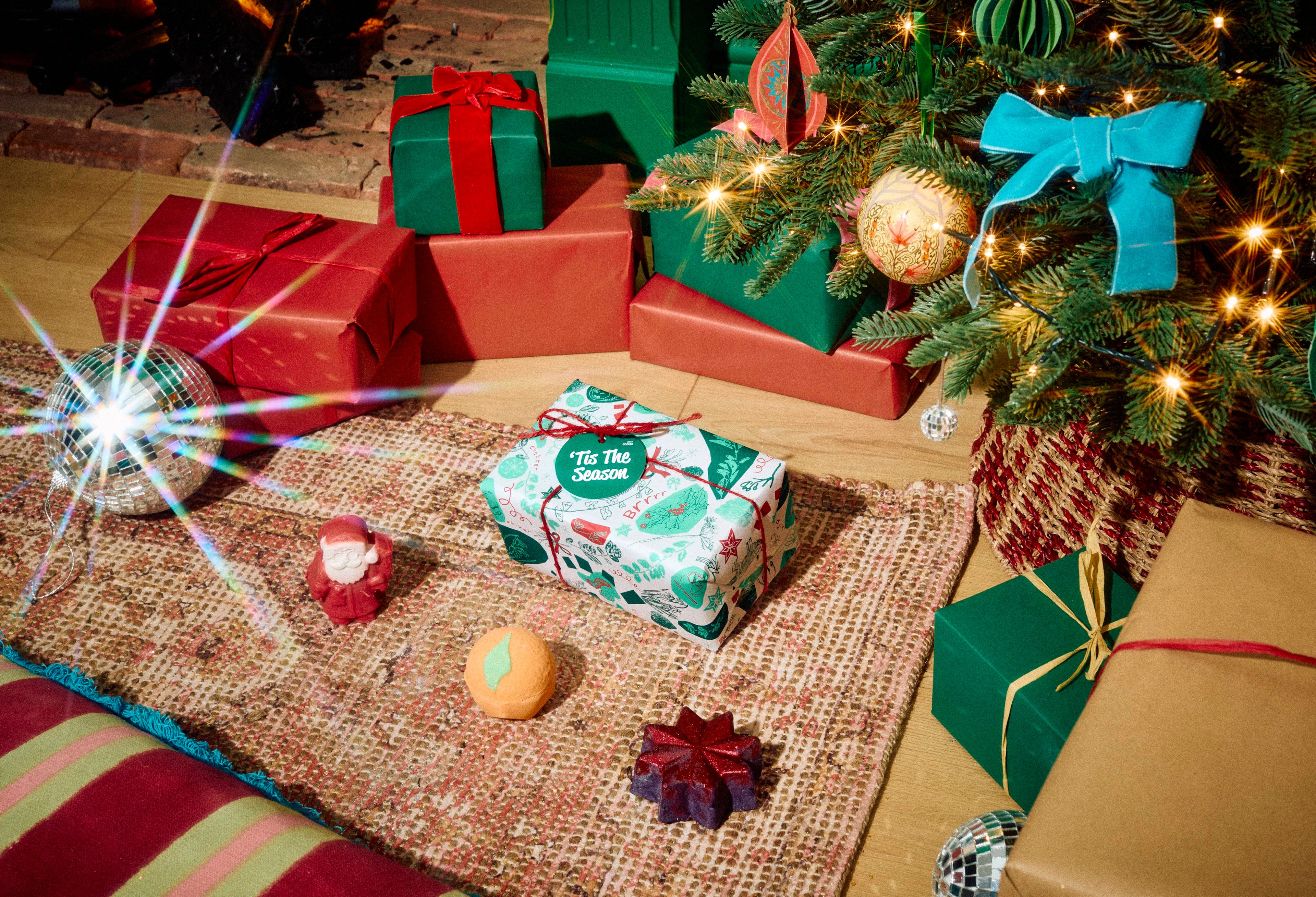 The Lush 'Tis The Season gift set rests on a patterned rug alongside an orange Clementine bath bomb, a star-shaped bubble bar, and a red Father Christmas soap, set in a festive room with wrapped presents and a Christmas tree.