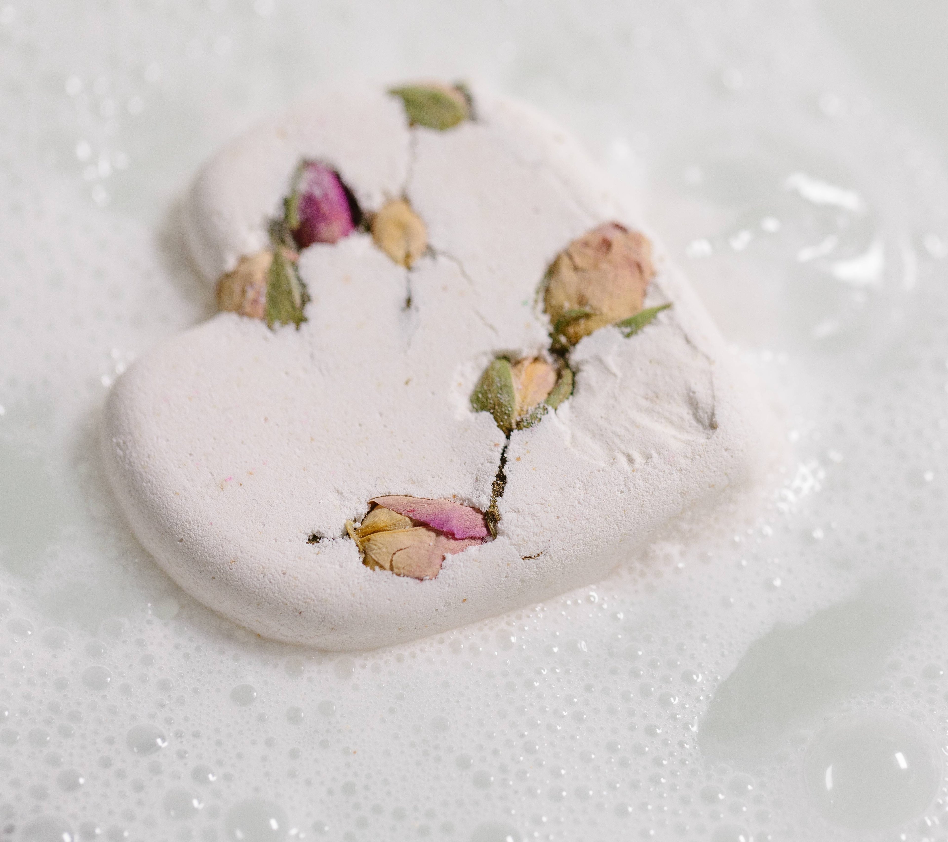 The image shows the Tisty Tosty bath bomb sitting on the water dispersing gentle white bubbles and dried rose buds throughout. 
