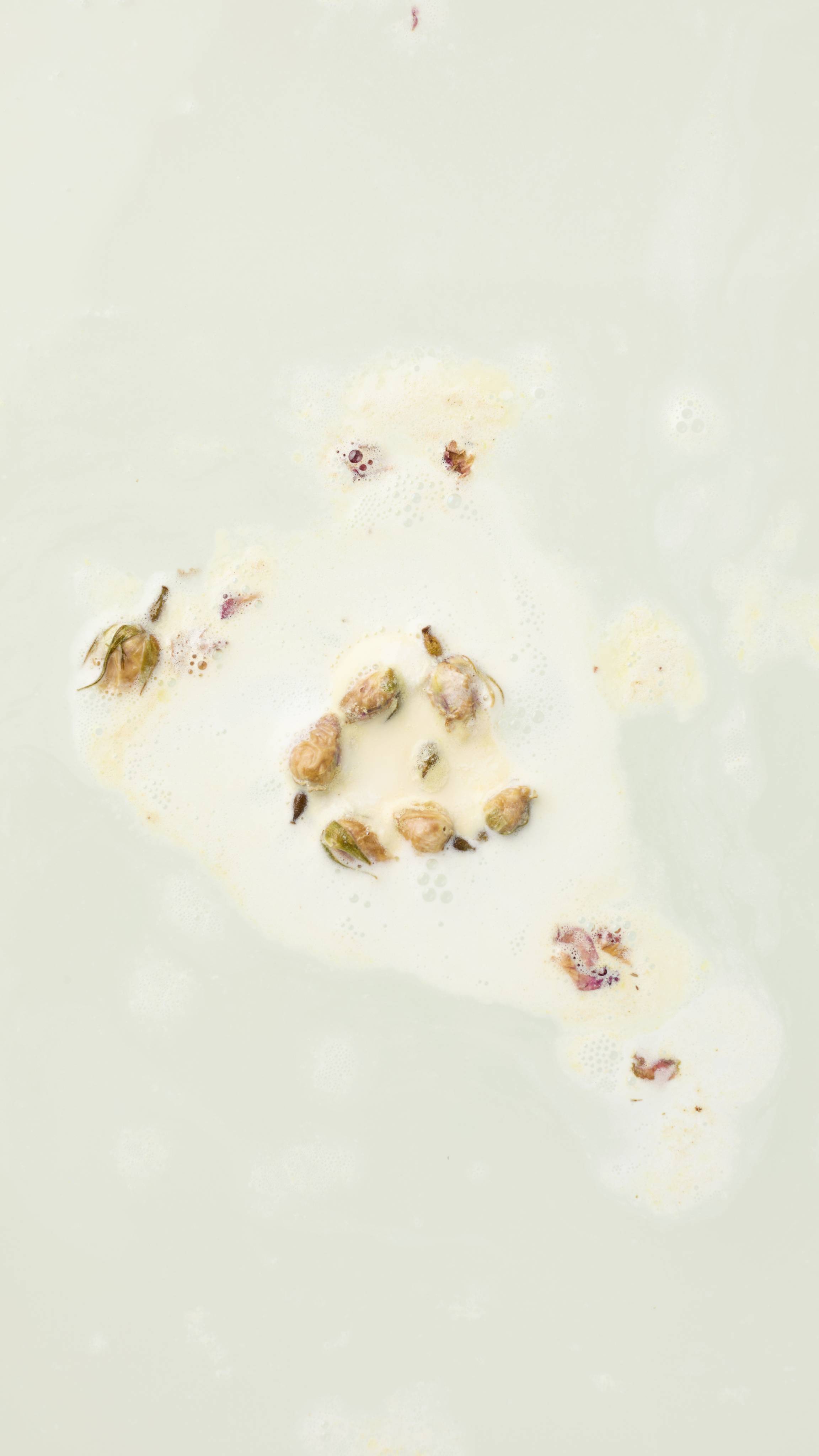 The image shows the Tisty Tosty bath melt almost fully dissolved into the milky water, leaving behind several whole, dried rose buds and smaller petal fragments. The buds float within a thinning patch of creamy white foam.