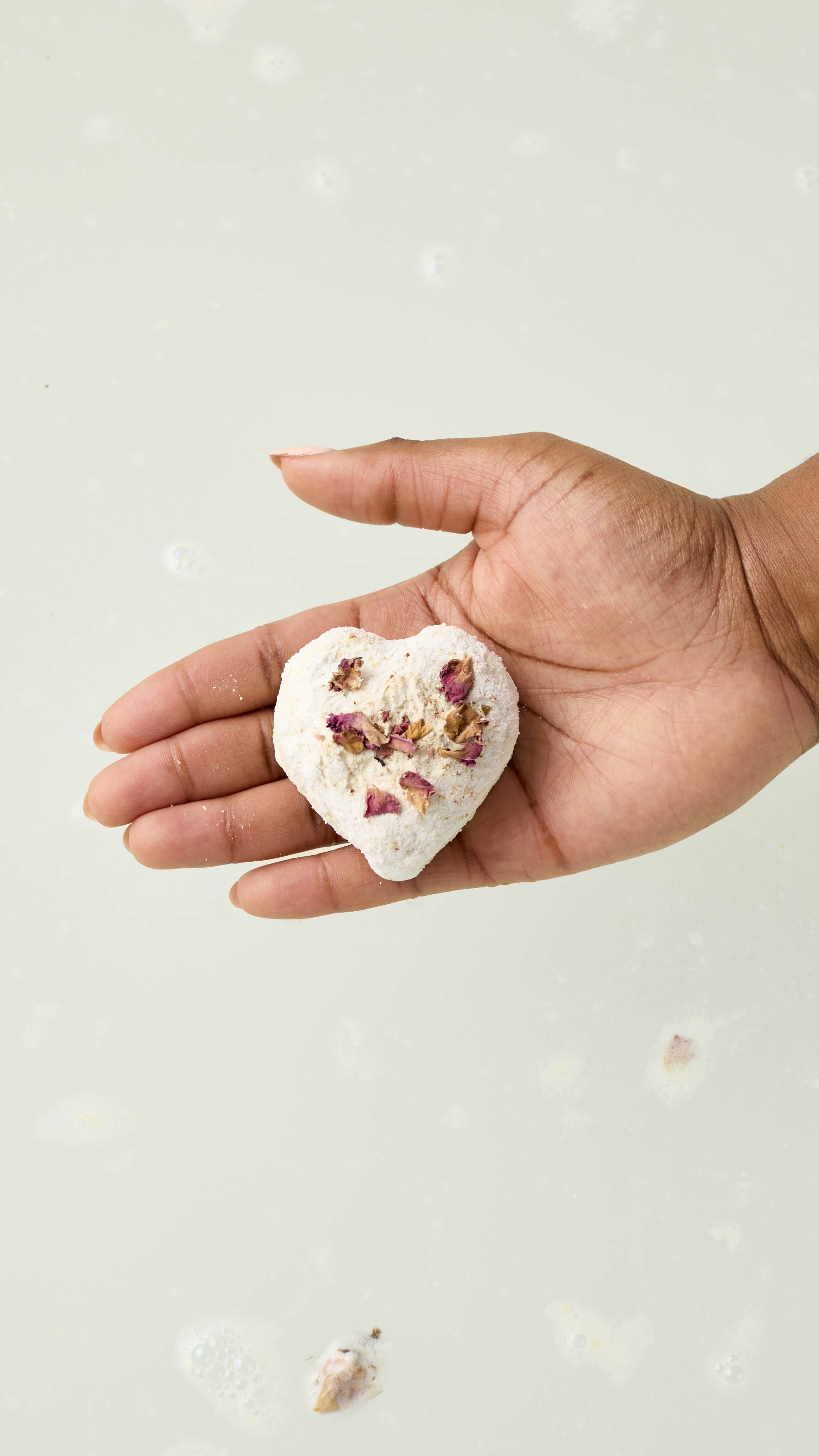 The image shows a hand holding the cream-coloured heart-shaped Tisty Tosty bath melt over a tub of milky, pale bathwater. Dried rose petals are visible on top of the bomb, and a few petals float on the surface of the water.