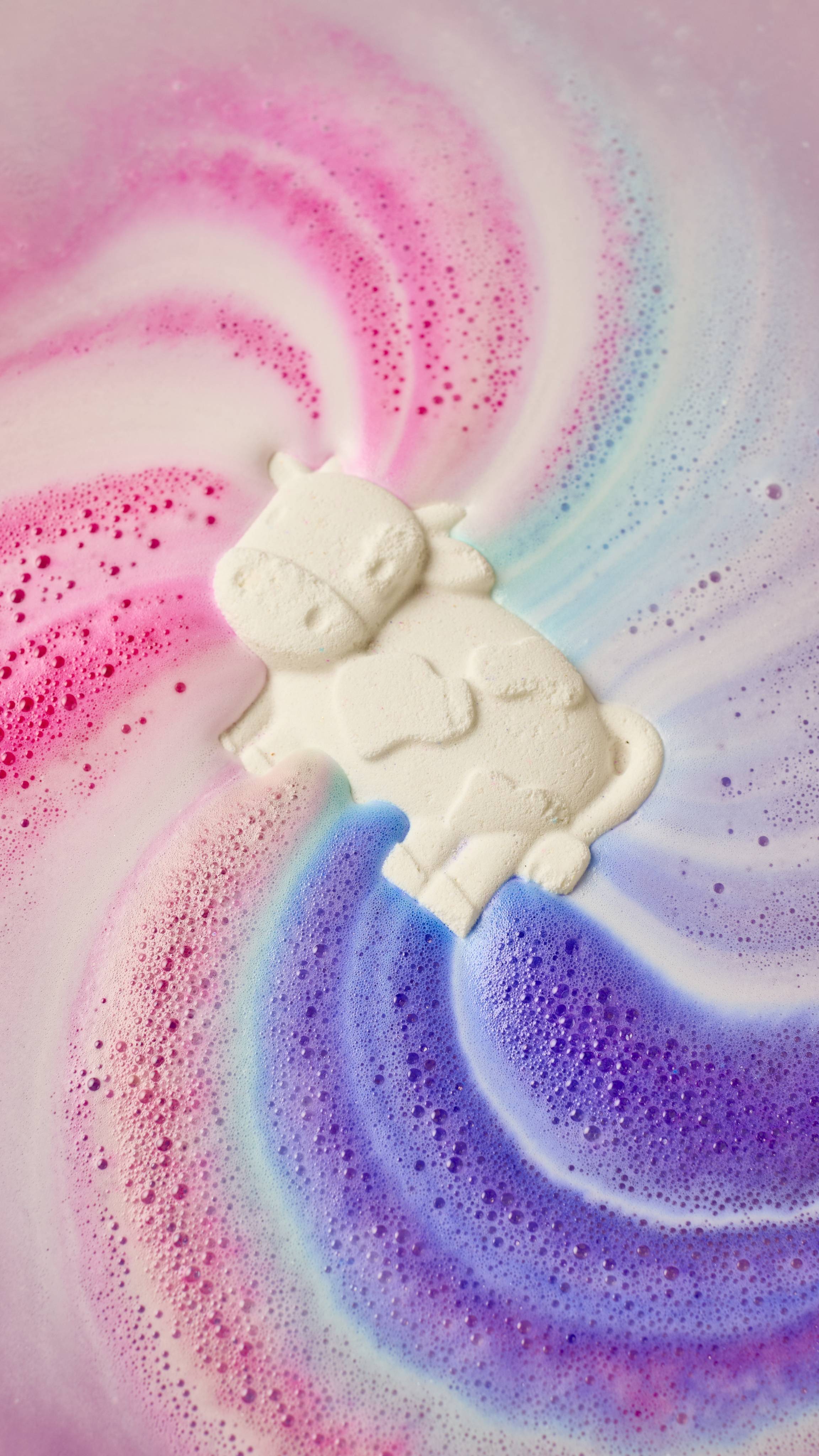 The Toby's Magic Cow bath bomb dissolves in water, releasing swirling trails of pink, purple, and blue foam. The white cow-shaped figure floats amidst the colourful, bubbly art.