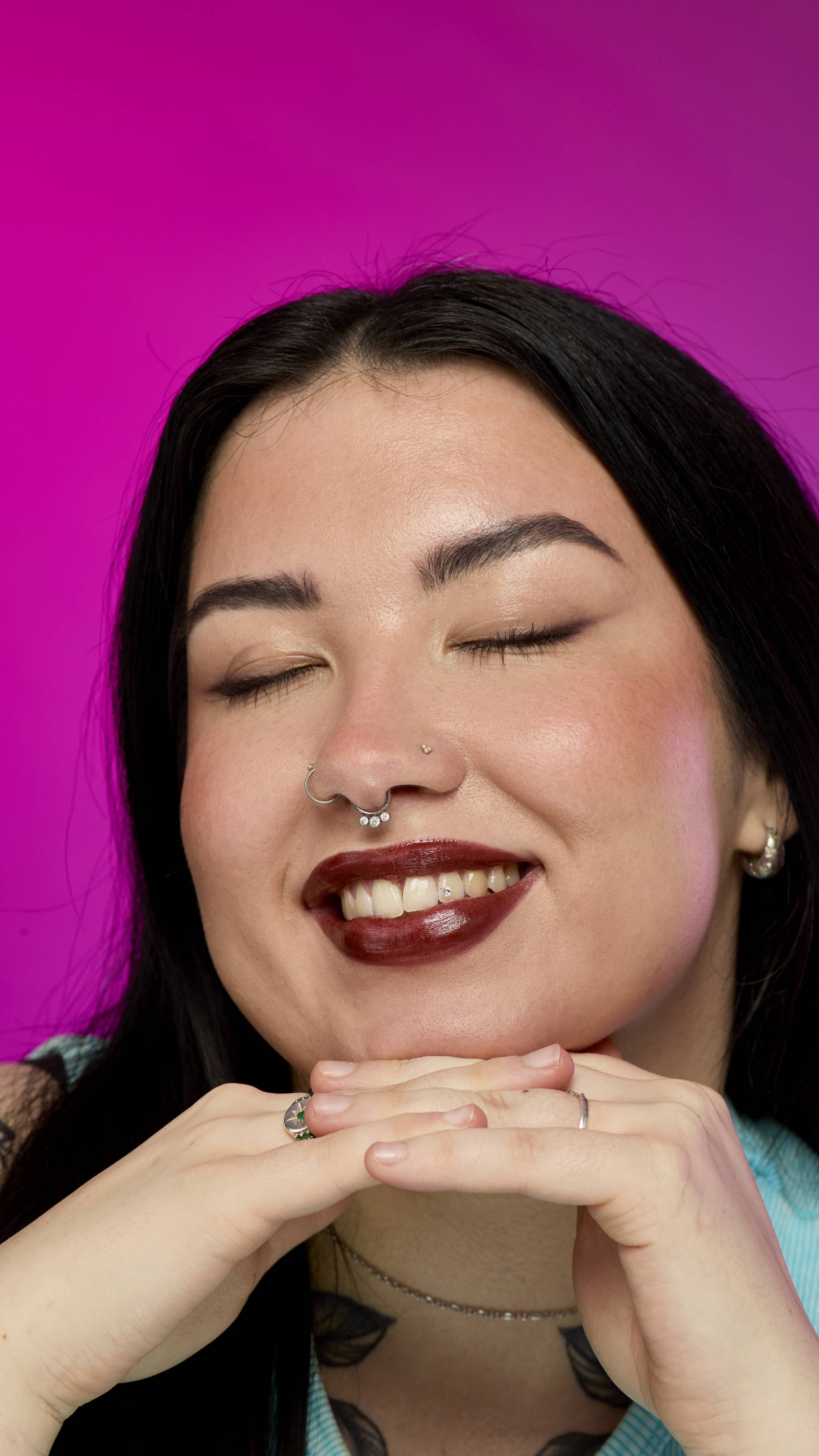 The image shows a model with long, dark hair on a vibrant magenta background, from the neck up, with their chin resting on interlocked hands. They are wearing the rich, dark chocolate-coloured Toffee Apple lipstick and smiling to reveal a tooth gem.