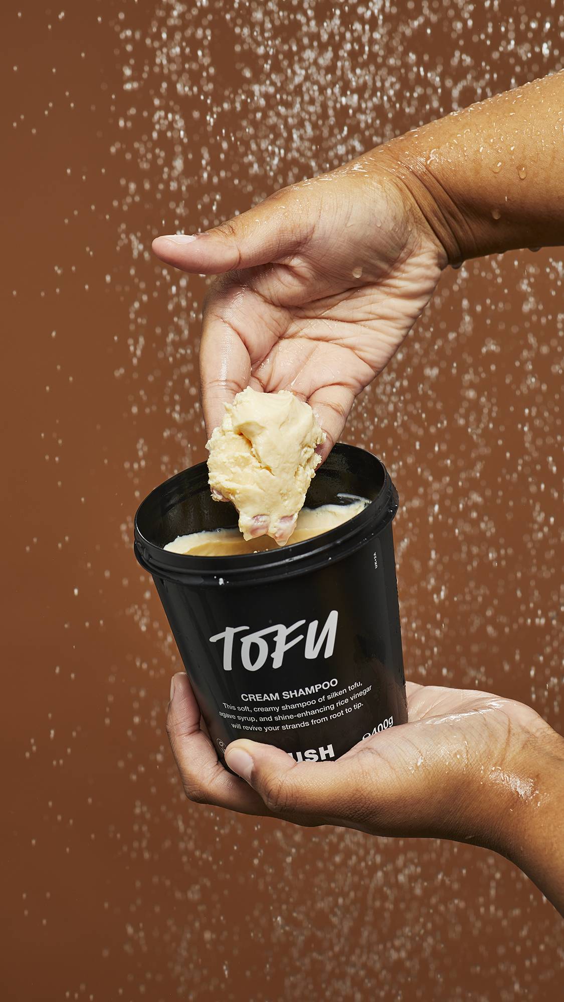 A close-up image of Tofu shampoo in a LUSH black pot under falling shower water as the model scoops out the product.