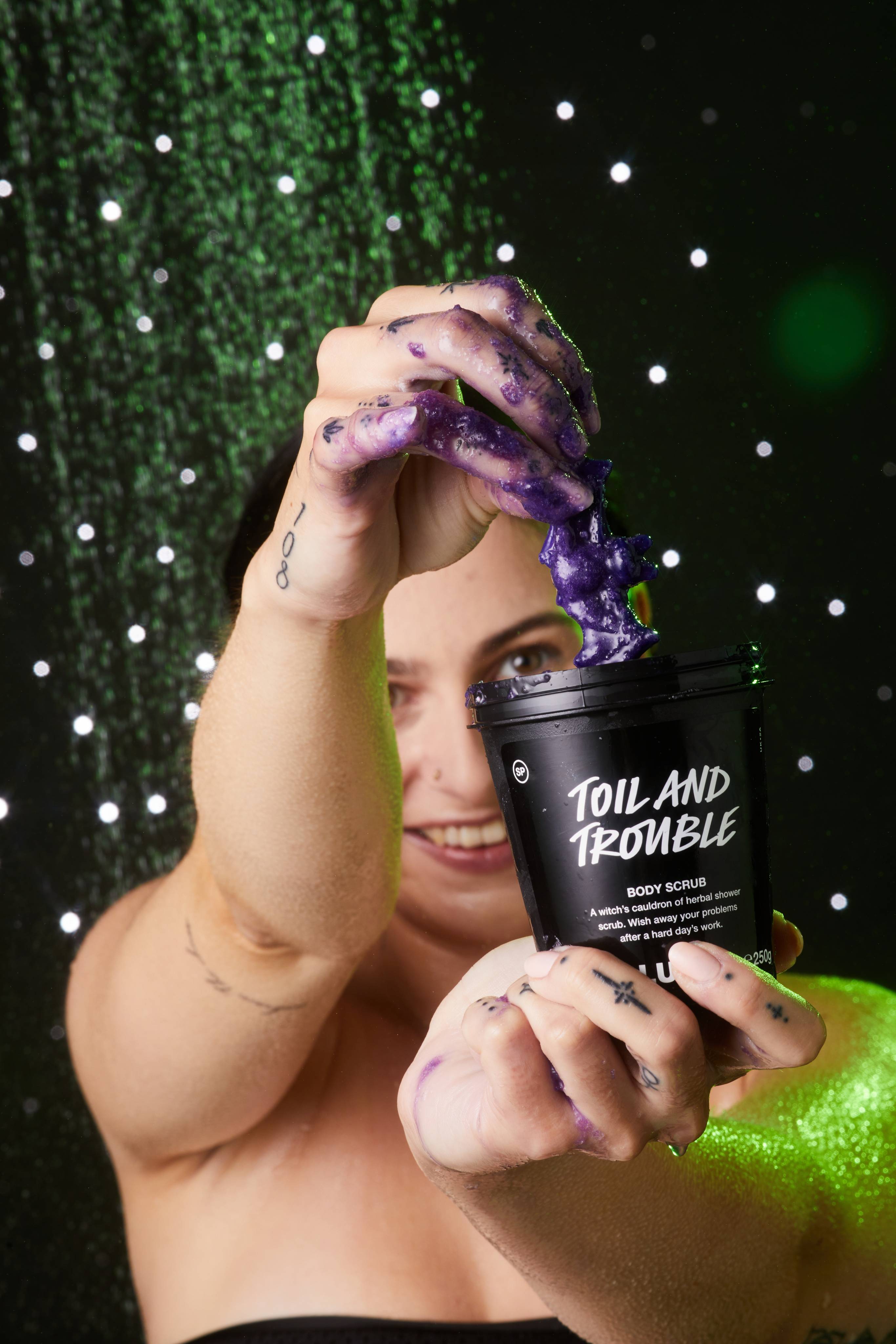 Model holds the product pot with one hand as they dip their other hand in, covering it in body scrub, pulling out the jelly bat.