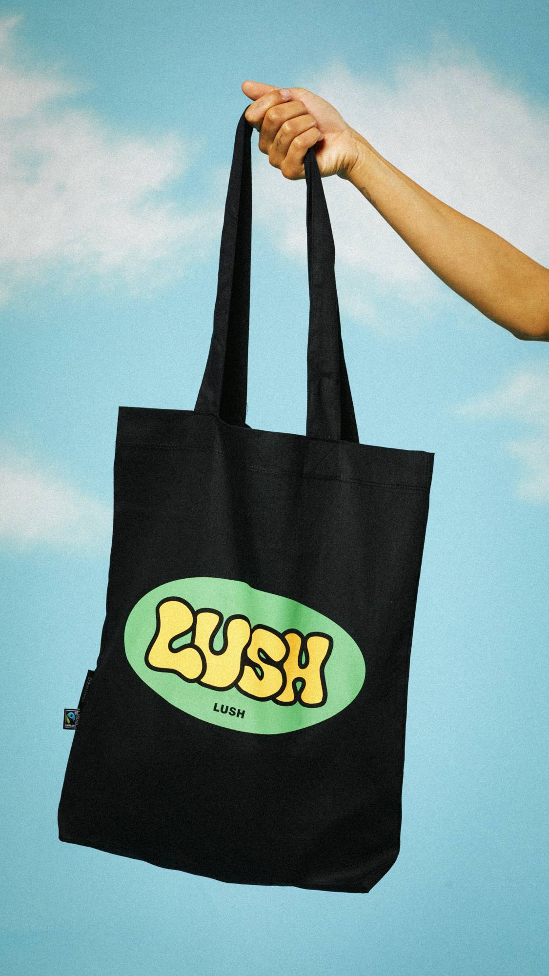 Image shows the model holding the Retro Bubble tote bag by the strings in the air on a blue sky background with clouds. 