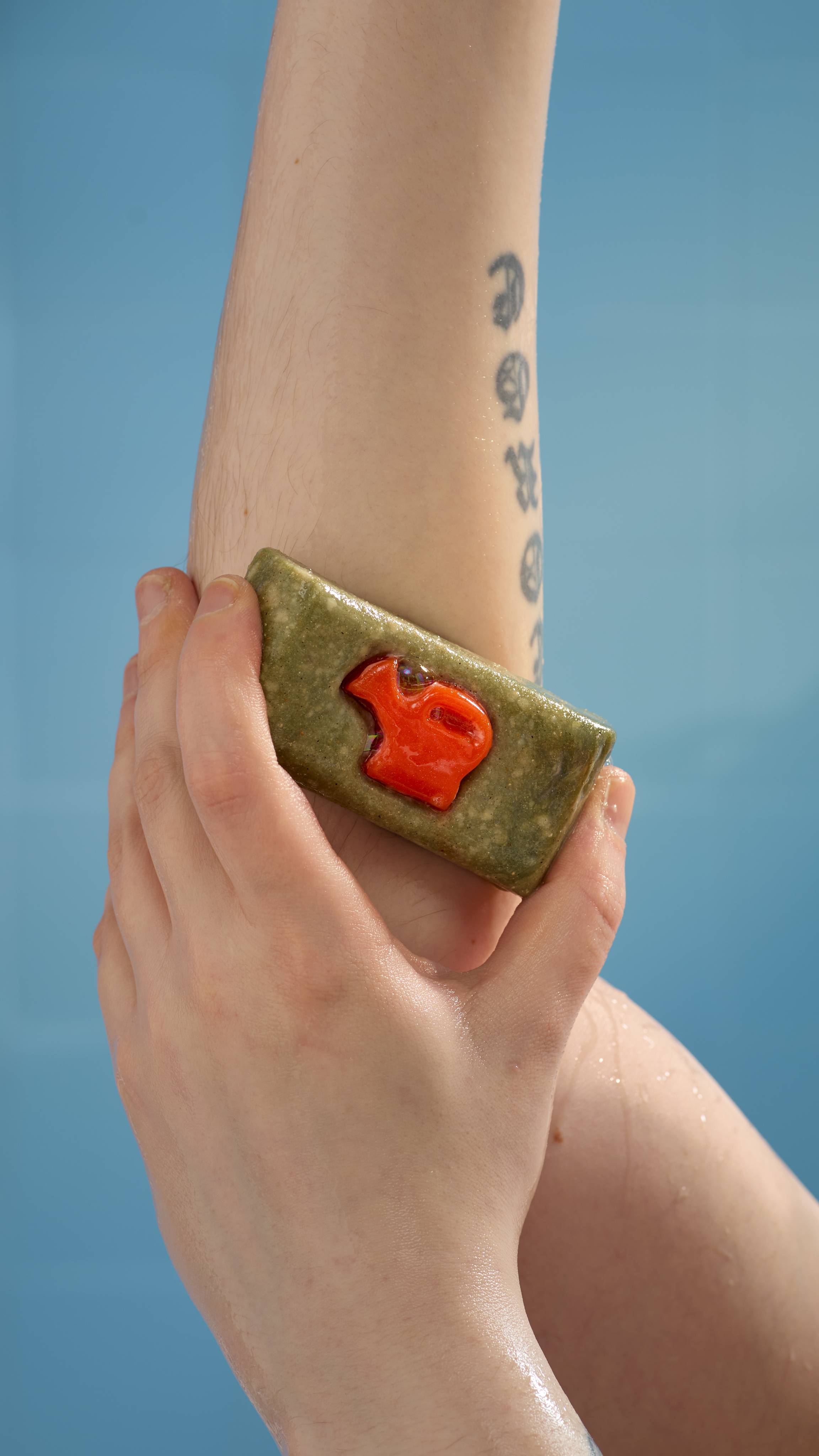 A close-up image of the model's forearm in the air are they lather up the True Grit soap over their elbow on a sky blue background. 