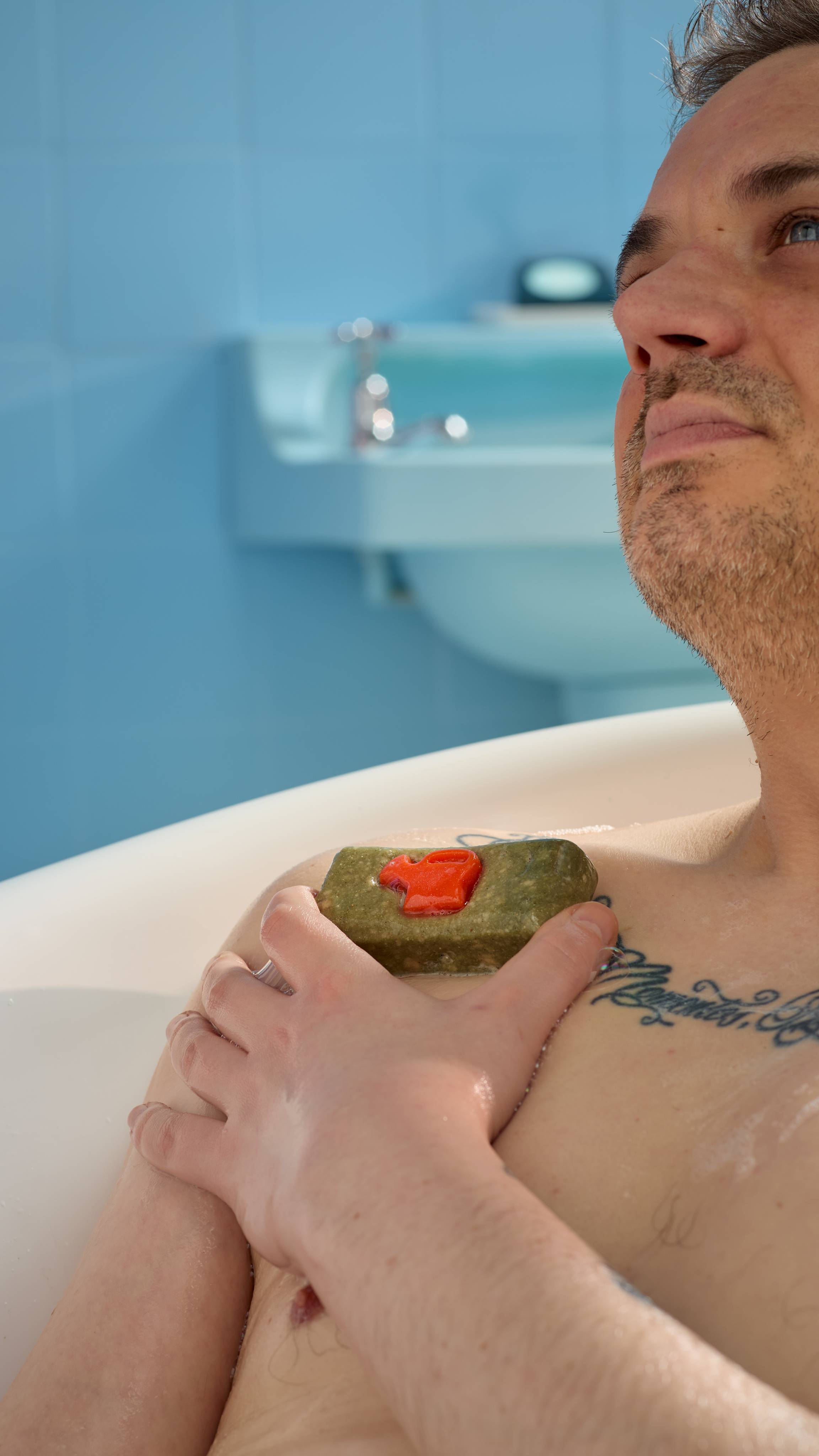 The image shows the model laid back in the roll-top bathtub as they lather up the True Grity soap over their shoulder. 