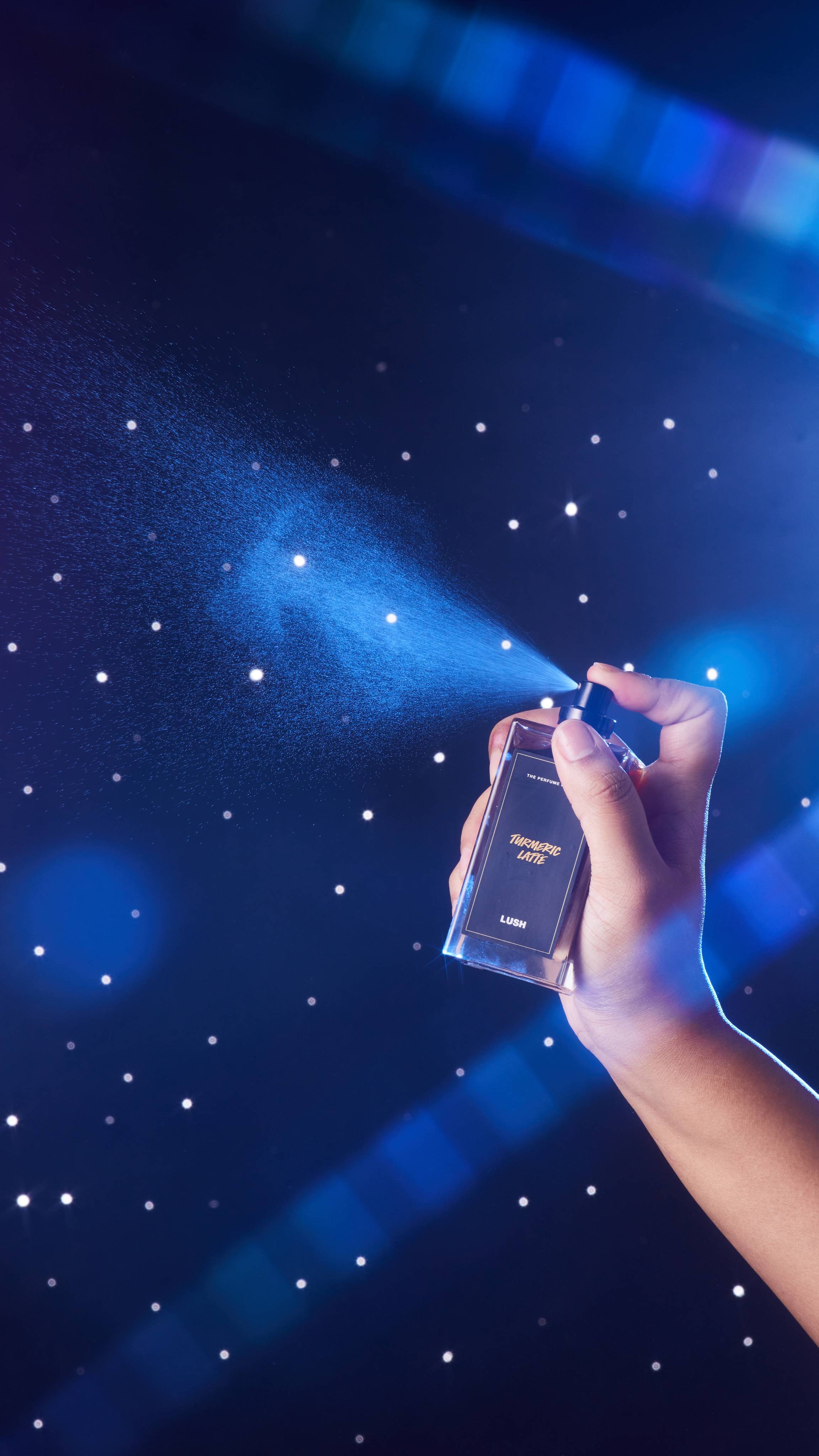 A close-up of the model's hand on a starry backdrop as they spritz the glass Turmeric Latter perfume bottle.