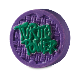 Turtle Power. A solid body scrub in the shape of a deep purple textured manhole cover with the words "TURTLE POWER" in green, graffiti-style writing on top.