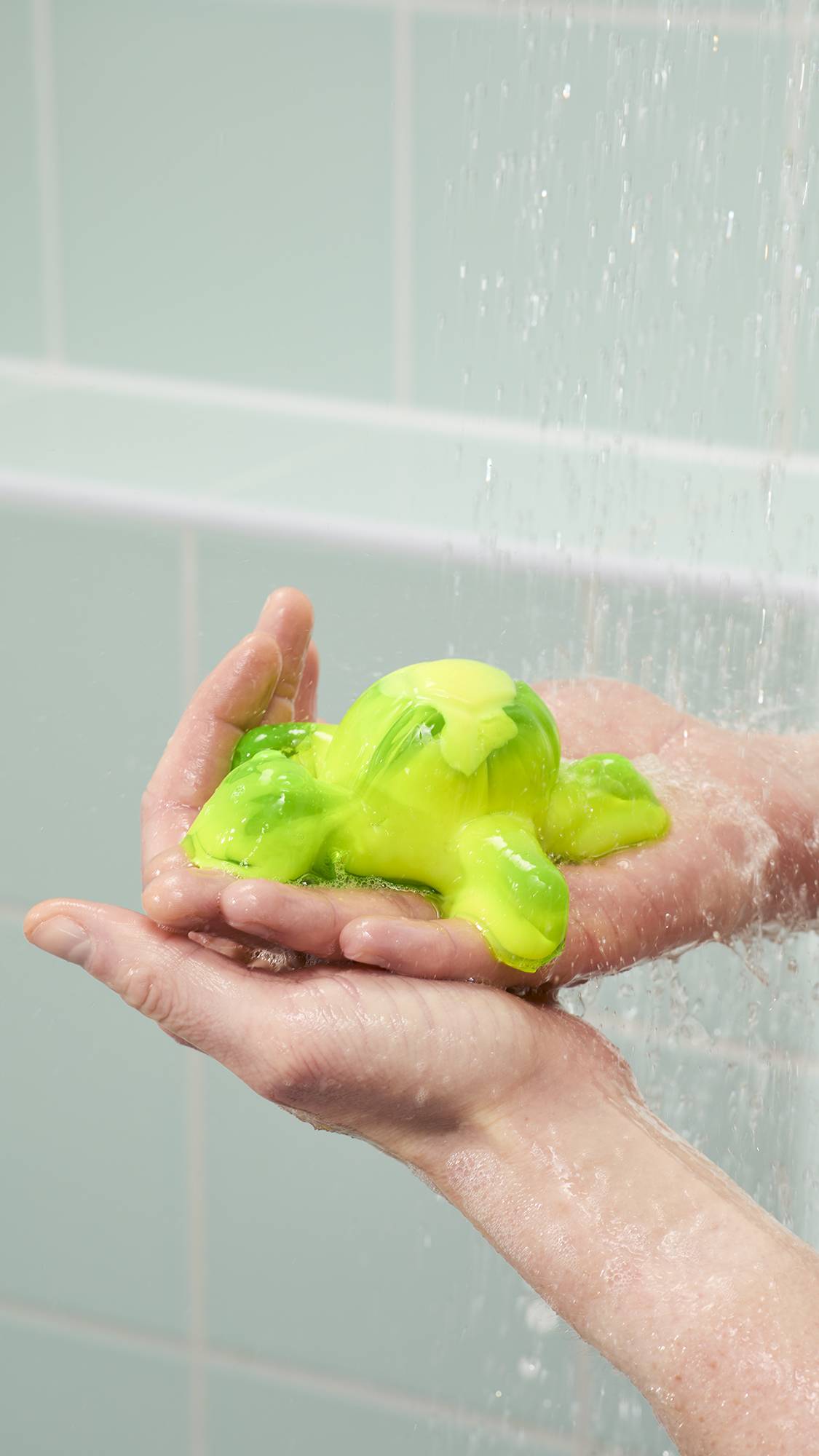 The image shows a vibrant lime-green and yellow shower jelly soap shaped like a sea turtle held in a person's cupped palms. The turtle is being rinsed under falling water in a shower against a backdrop of pale green tiles.