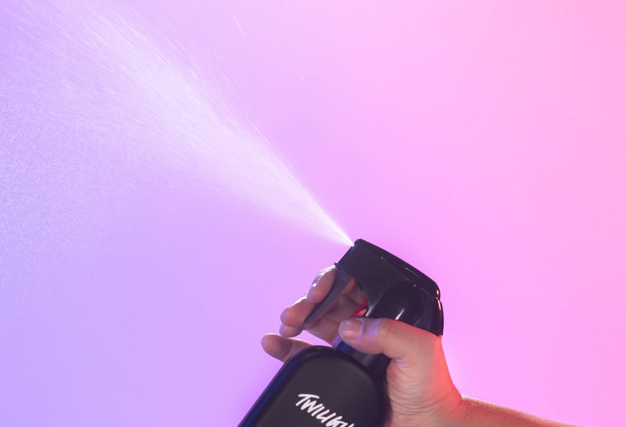 Twilight body spray is sprayed up into the air, in front of a bright, light purple to light pink gradient background.