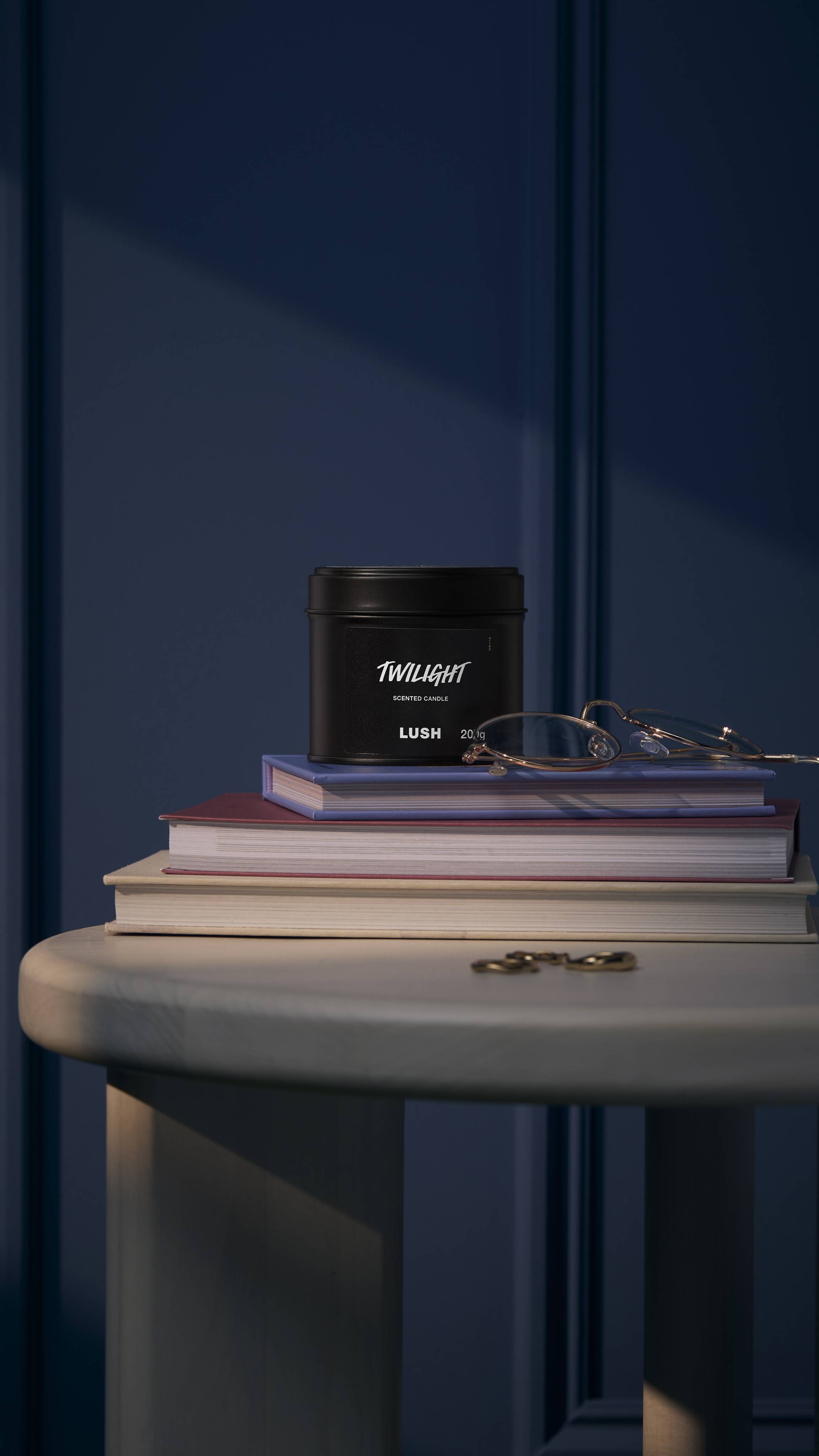 The image shows a deep, midnight-blue walled background. The black tin Twilight candle sits on a white, wooden side table on top of a small pile of books, glasses, and jewellery. 