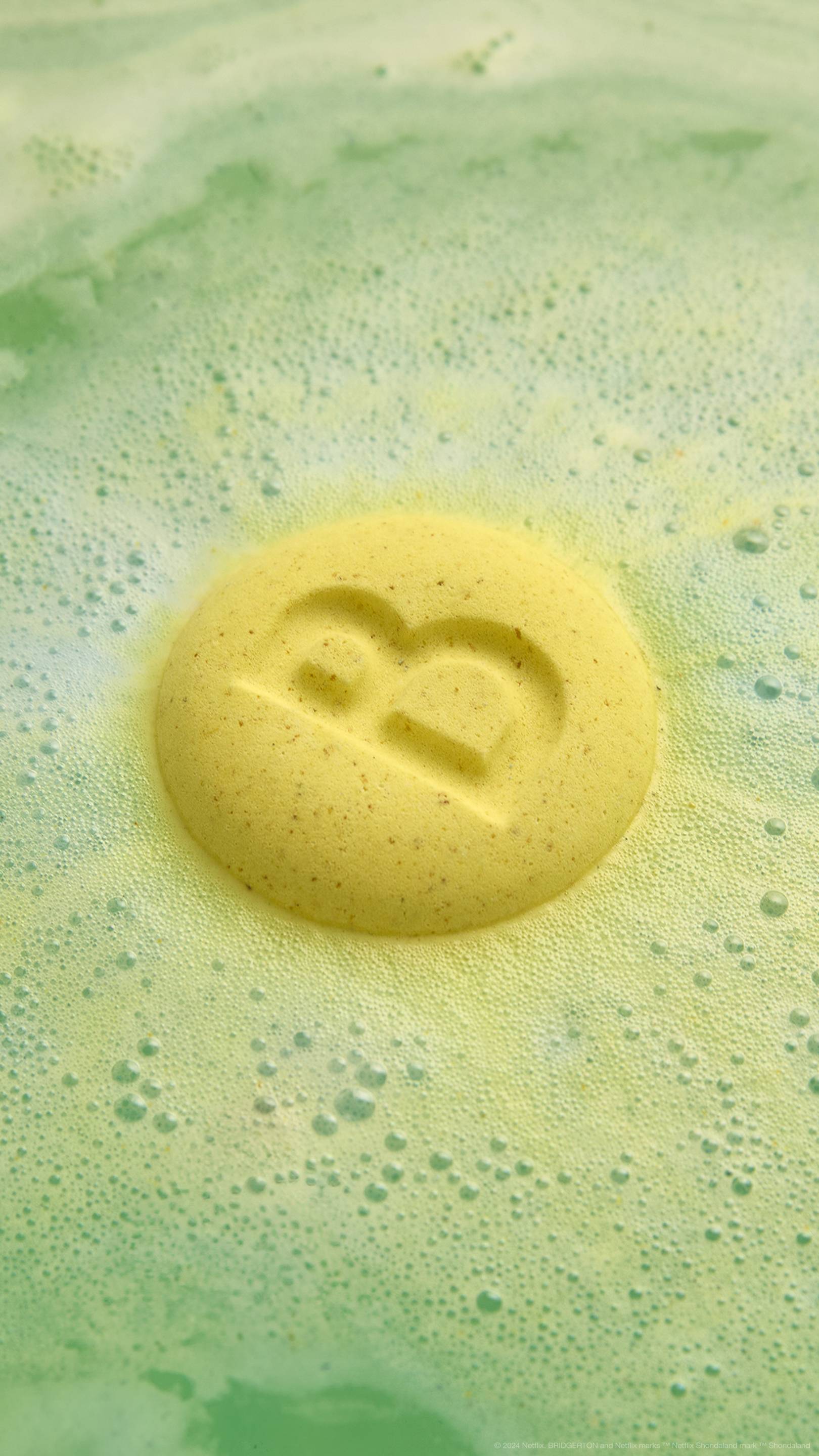 The image shows the Two Families bath bomb on the surface of the water with the yellow side with an embossed letter "B" facing up. Fresh green water lies beneath a layer of velvety foam.