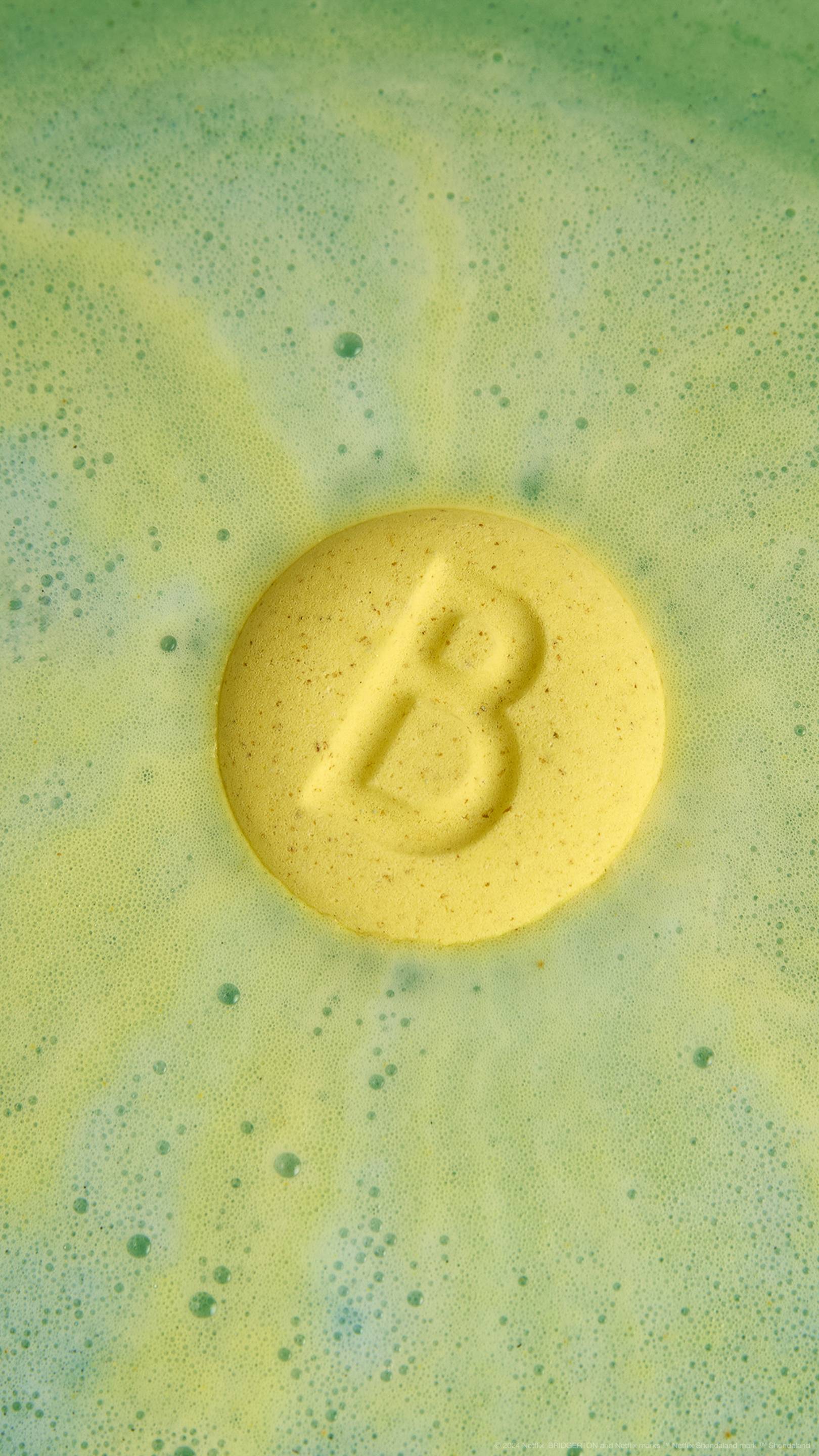 The image shows the Two Families bath bomb on the surface of the water with the yellow side with an embossed letter "B" facing up. Fresh green water lies beneath a layer of velvety foam.