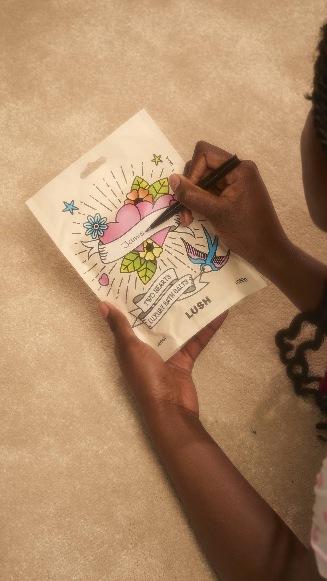 A person uses a pen to write the name ‘Jamie’ on the banner of a packet of Lush Two Hearts Luxury Bath Salts. The packet features a colourful heart and floral tattoo-style design, set against a soft, neutral carpet background.