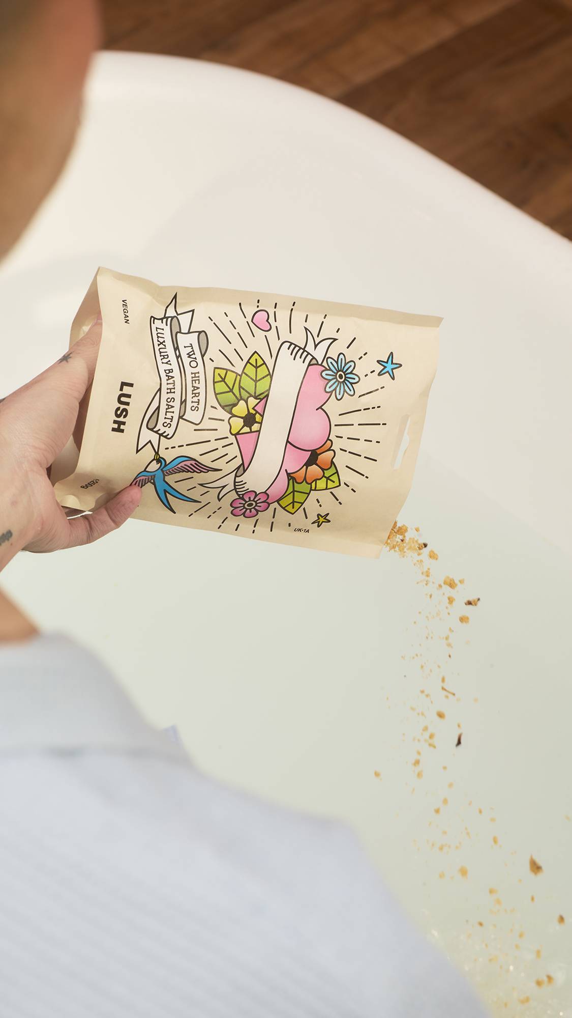 A hand pours Lush "Two Hearts" luxury bath salts into a bathtub of water. The cream-coloured packet features a central banner surrounded by colourful, tattoo-style illustrations of hearts, flowers, and a swallow.