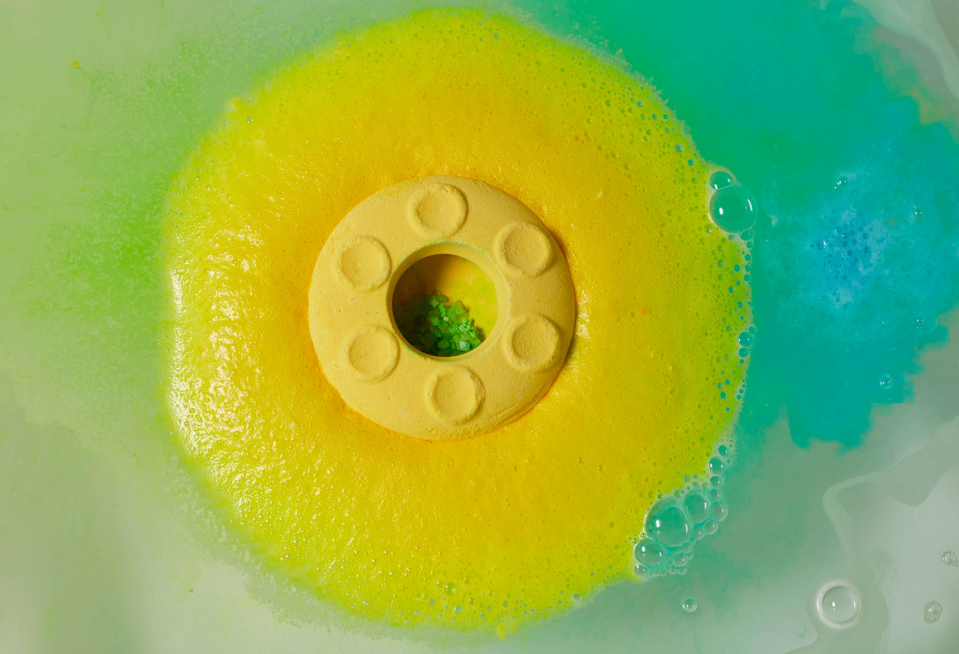 The circular UFO bath bomb floats on the water surrounded by yellow foam. Inside the hollow bath bomb are green salt crystals.