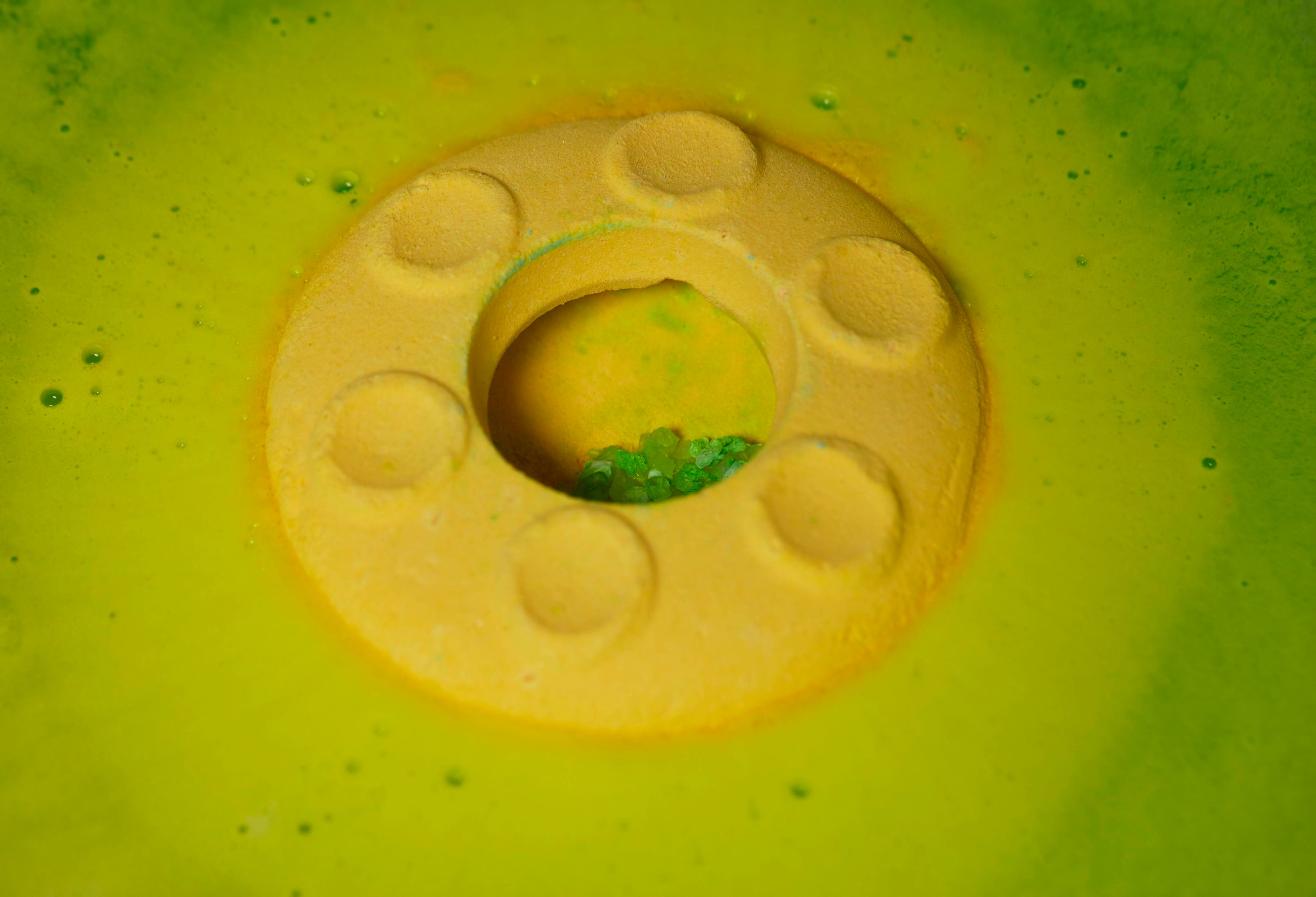The yellow bath bomb mixes with the blue top on a silky sea of green foam and shows the green salt crystals inside.
