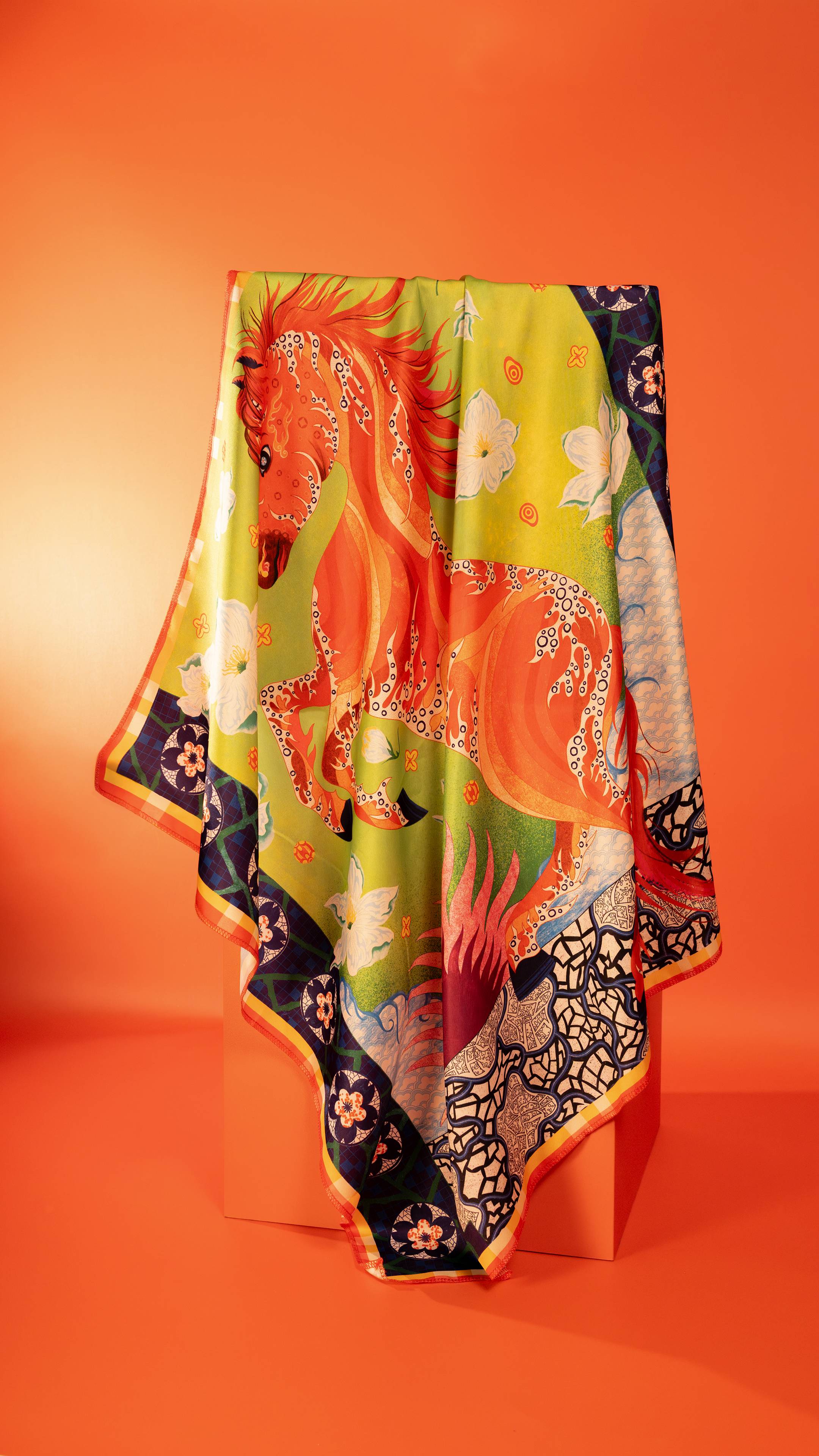 The Untamed Flame Knot Wrap drapes over a display, showcasing a vibrant illustration of a rearing red horse on a lime green background. The fabric is bordered by blue geometric patterns and white flowers, set against a solid orange backdrop.