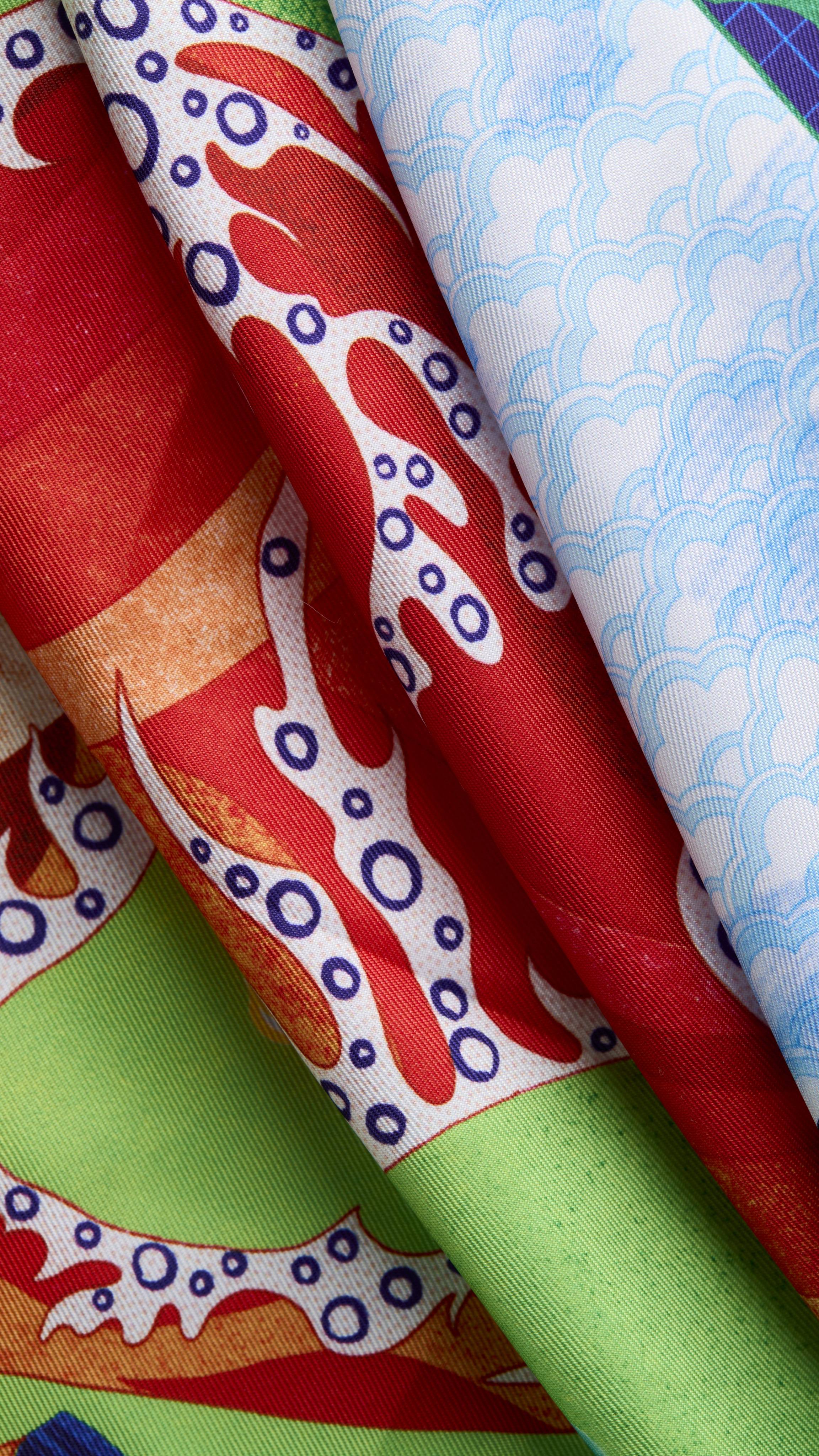 A close-up of the folded Untamed Flame Knot Wrap reveals vibrant red and orange wave patterns dotted with white circles, layered against bright green sections and soft blue cloud illustrations.
