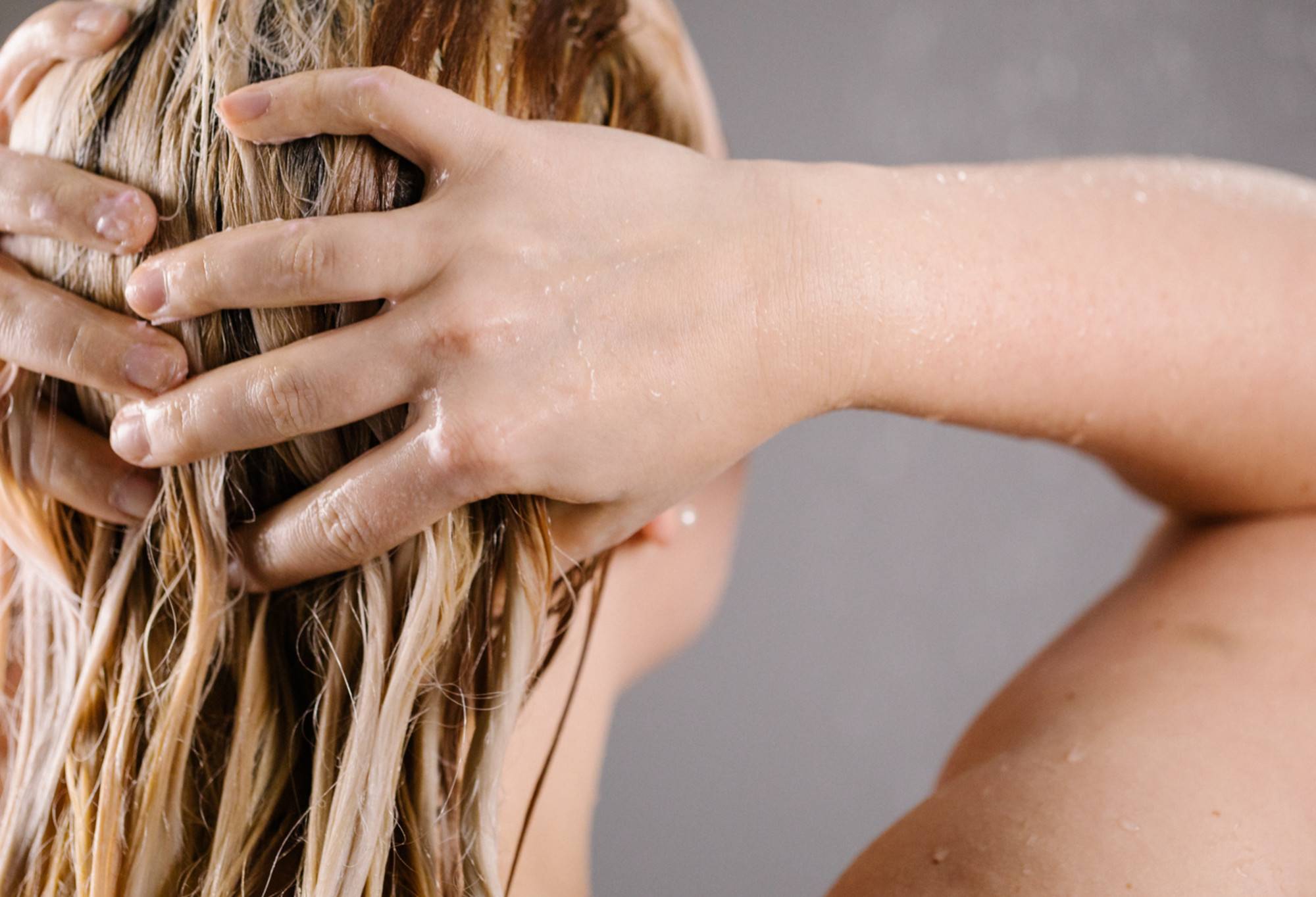 A person with long, blonde hair, coated in light pink Valkyrie conditioner, massages the back of their head with both hands.