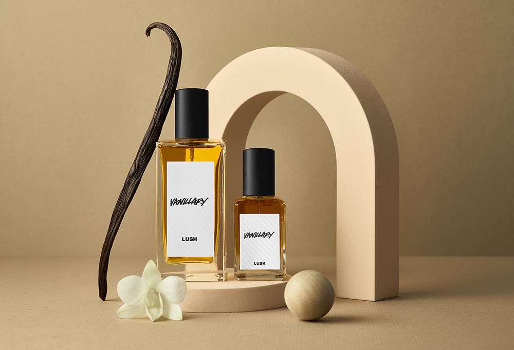 Vanillary perfume sits upon a cream podium and semi circle. A Vanilla pod and small wooden ball sit against the fragrance.