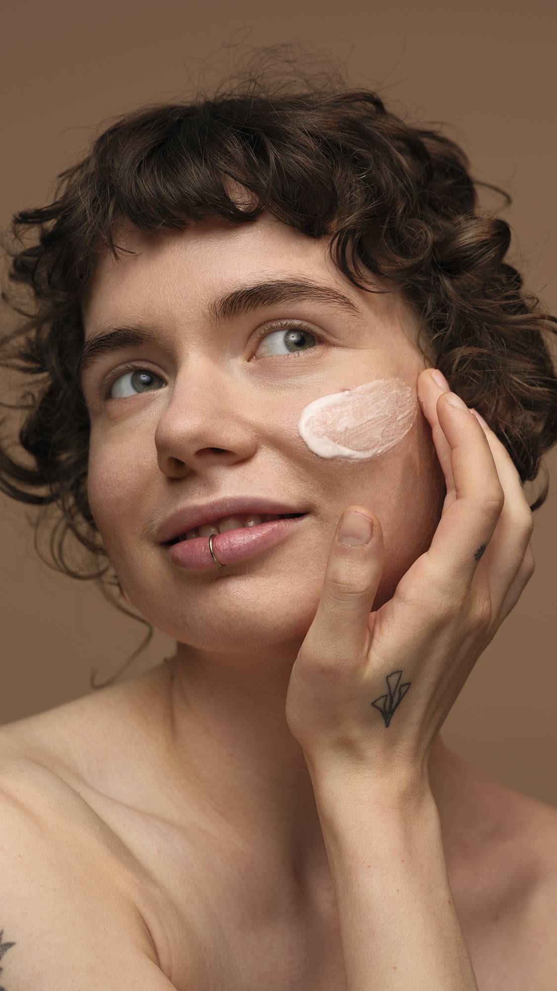 The image shows the model with curly brown hair on a warm, earth-toned background as they gently smooth the Vanishing Cream self-preserving moisturiser over their cheek. 