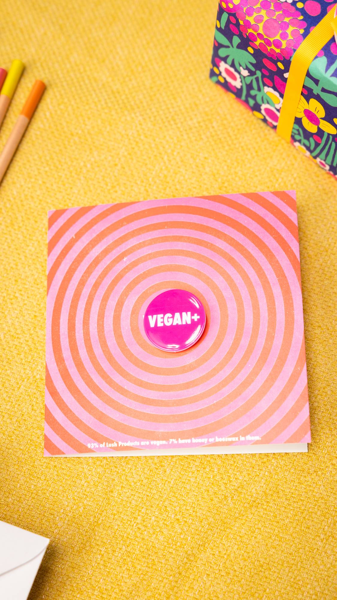 Image shows the Vegan + greetings card on a yellow cloth - the pink badge sits prominently in the centre of the card.