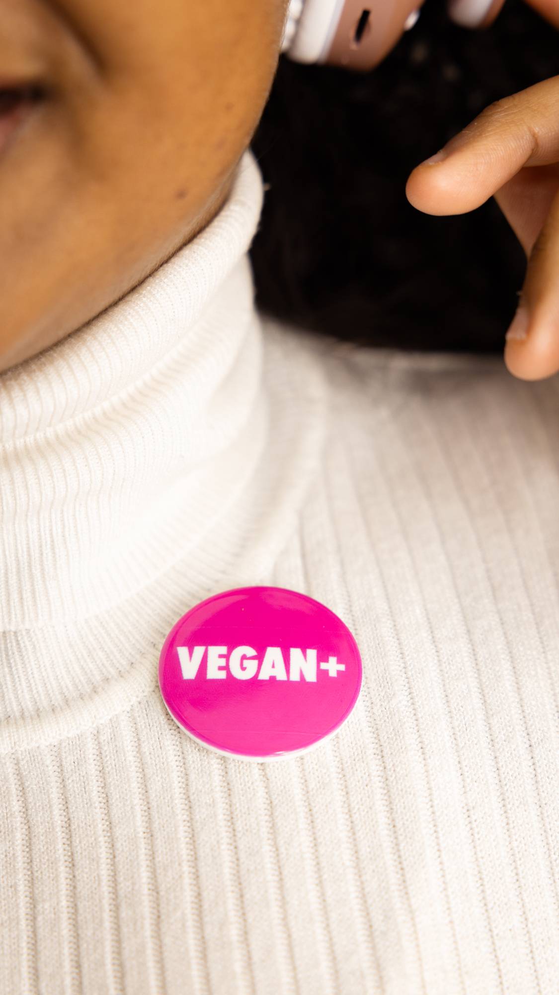 Image shows a super close-up of model wearing the bright pink VEGAN + pin badge on a white turtleneck jumper. 