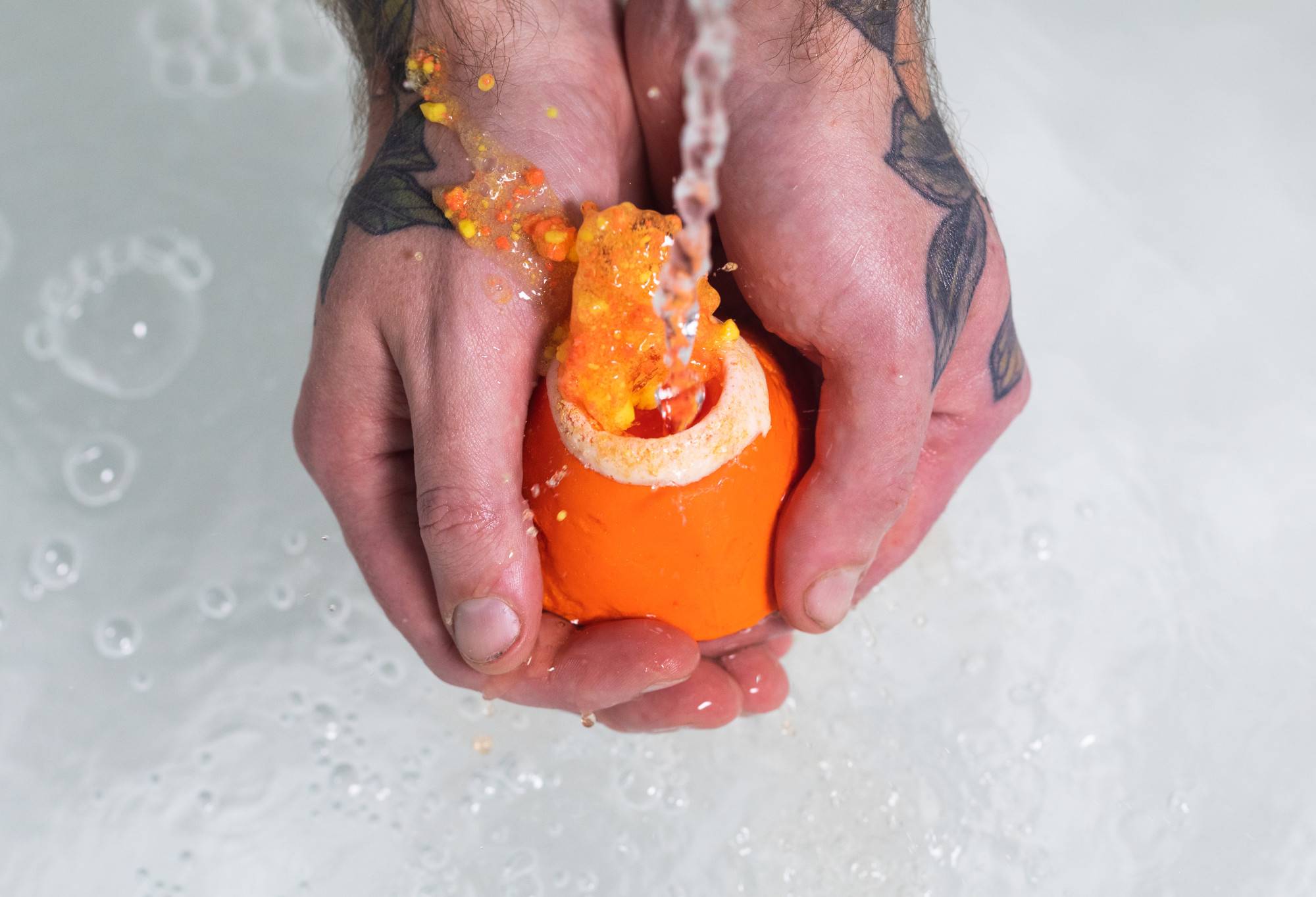Two hands with tattoos cup Vesuvio. Water runs in the open top. Orange water spurts out leaving specks of orange on the hands.