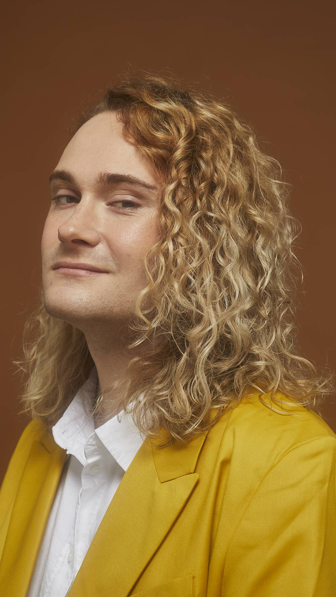 Model smiles and wears a bright yellow jacket showing defined, golden, curled locks on an earth-toned background.