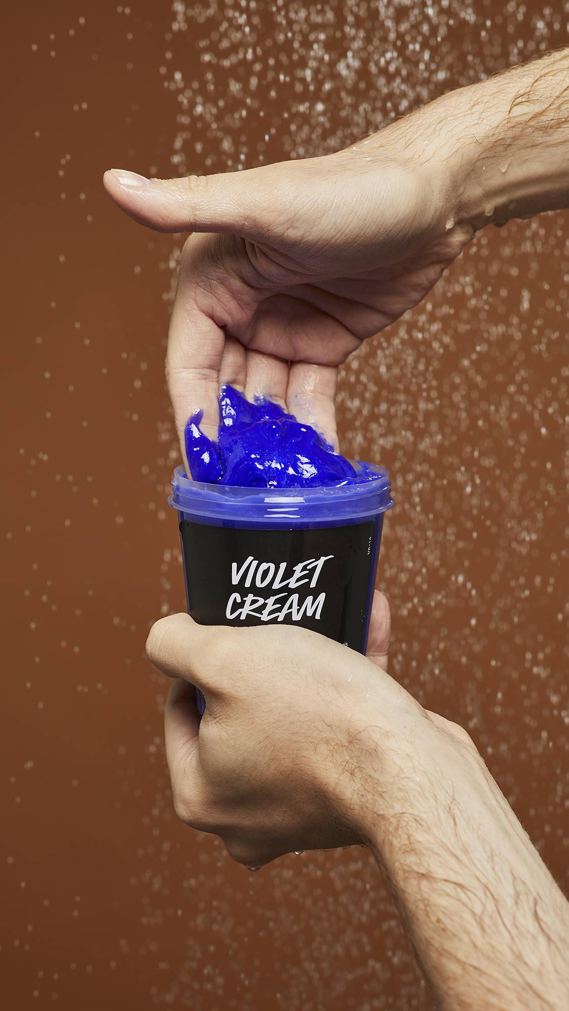 A close-up image of model scooping the vibrant Violet Cream shampoo out of the LUSH pot under running shower water.