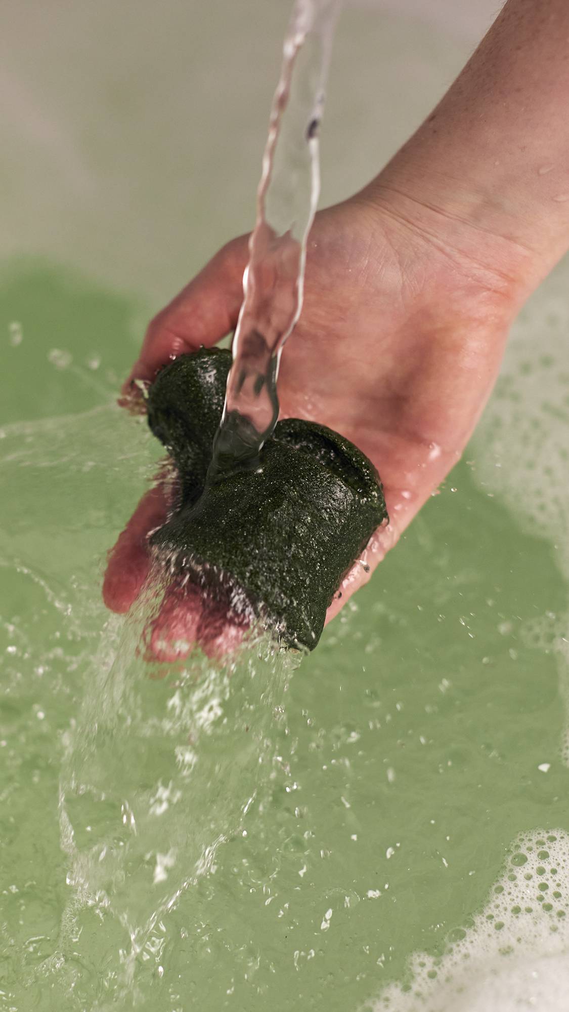 The image shows a close-up of the model holding the Wash And Grow bubble bar under running water making bubbles below. 