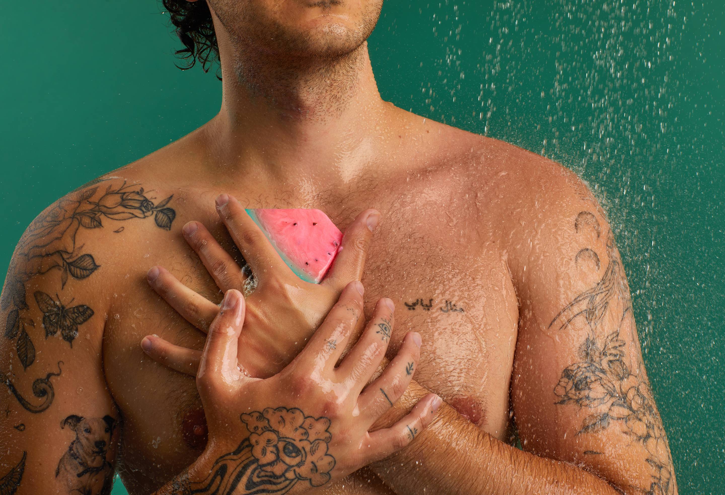 The image shows a person holding a slice of Watermelon Soap against their chest. The triangular soap has layers of pink and green. Water droplets are visible on the skin and product, against a dark teal background.