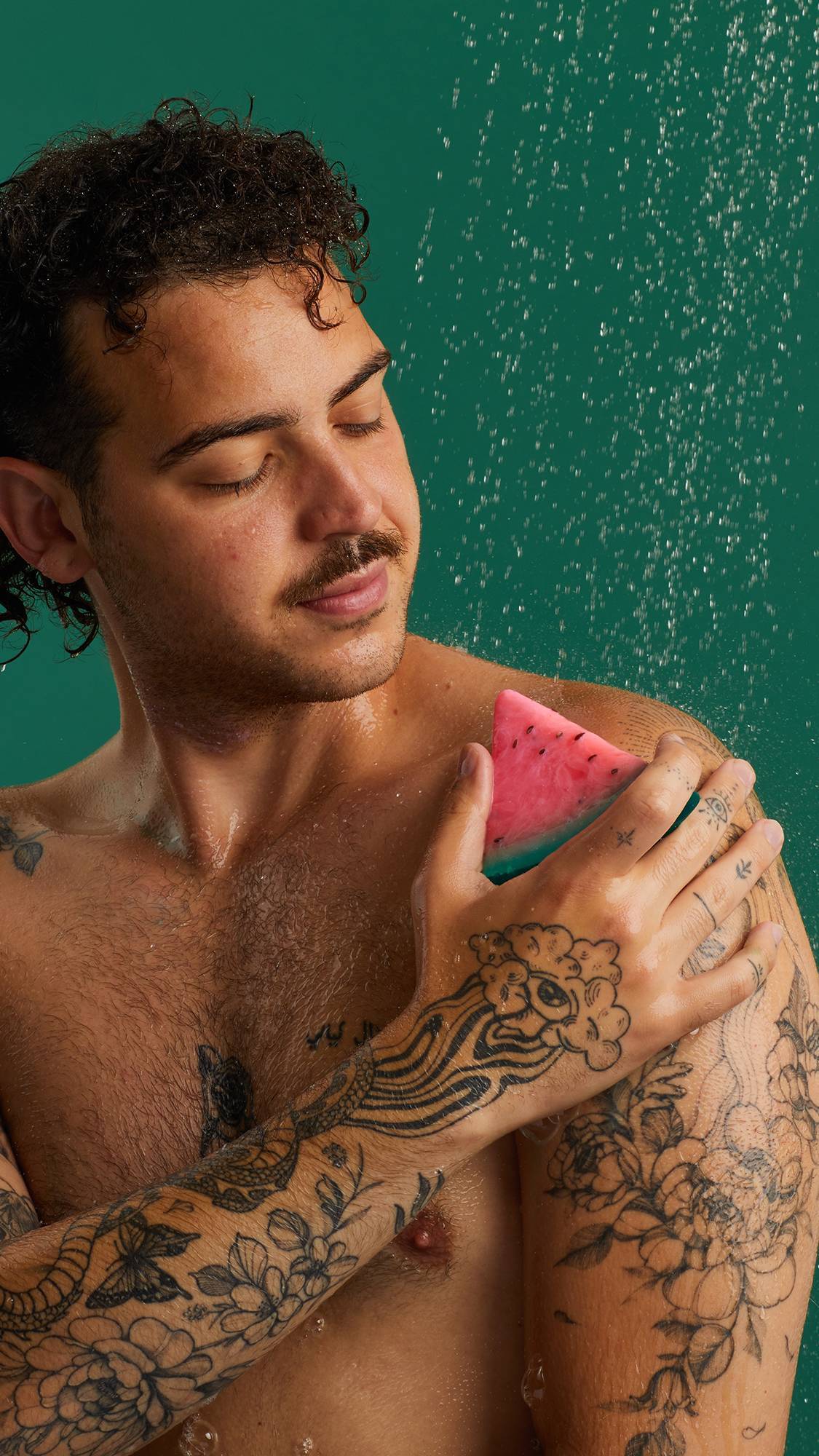 The image shows a person with a serene expression, rubbing a slice of Watermelon Soap on their tattooed shoulder. The triangular product has pink and green layers. Water droplets fall on their skin against a dark teal background.