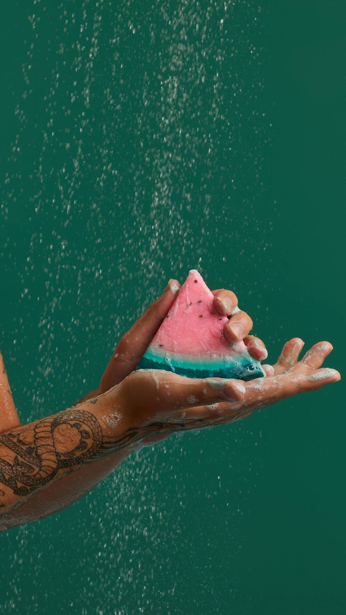 The image shows hands holding a slice of 'Watermelon' soap. The product, with pink "flesh", green outer layers and small black seeds, rests in a cupped palm with visible lather. Water droplets fall against a dark teal background.