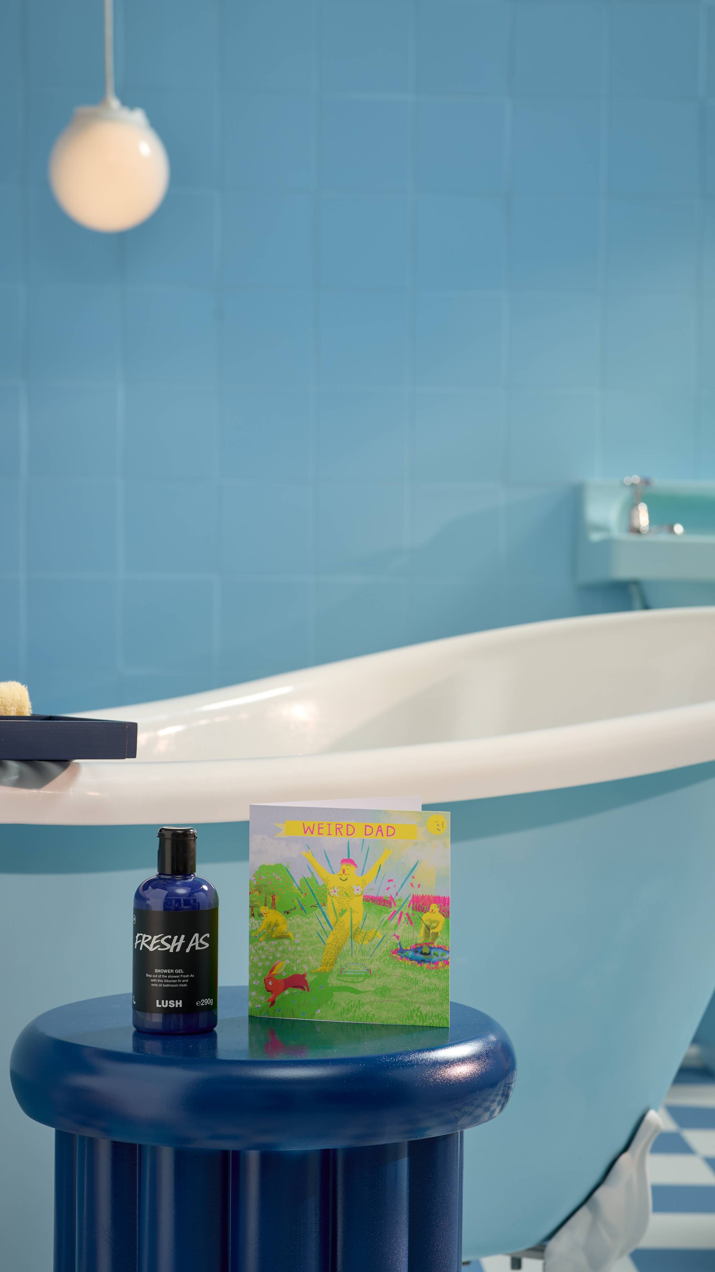 The image shows a blue, tiled bathroom with a roll-top bathtub. There is a bright blue stool in front of the bath with the Fresh As shower gel and Weird Dad card on top. 