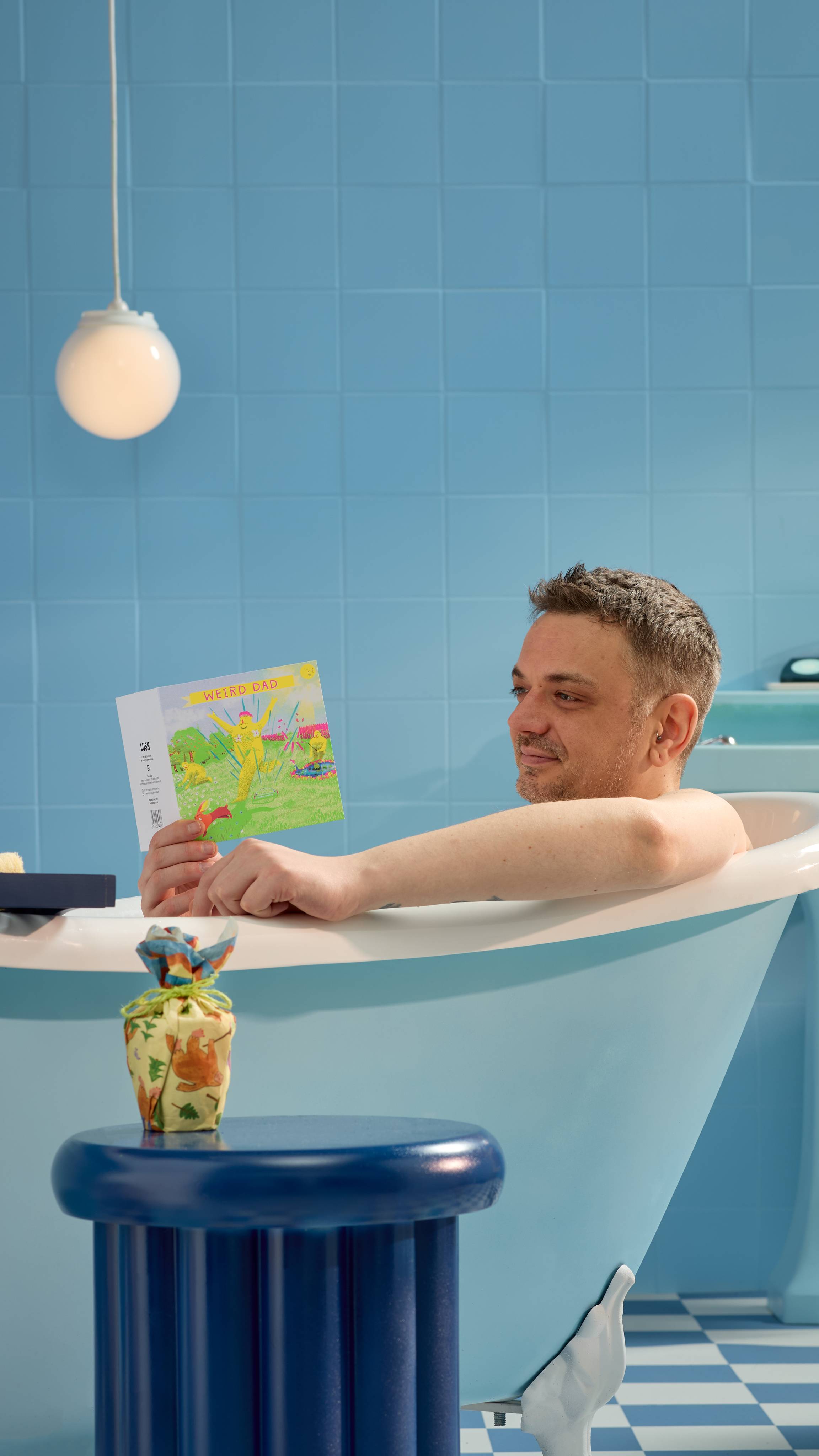 The model is sitting in a roll-top bathtub in a blue, tiled room reading the Weird Dad gift card as a wrapped gift is sat on a blue stool beside the bath. 