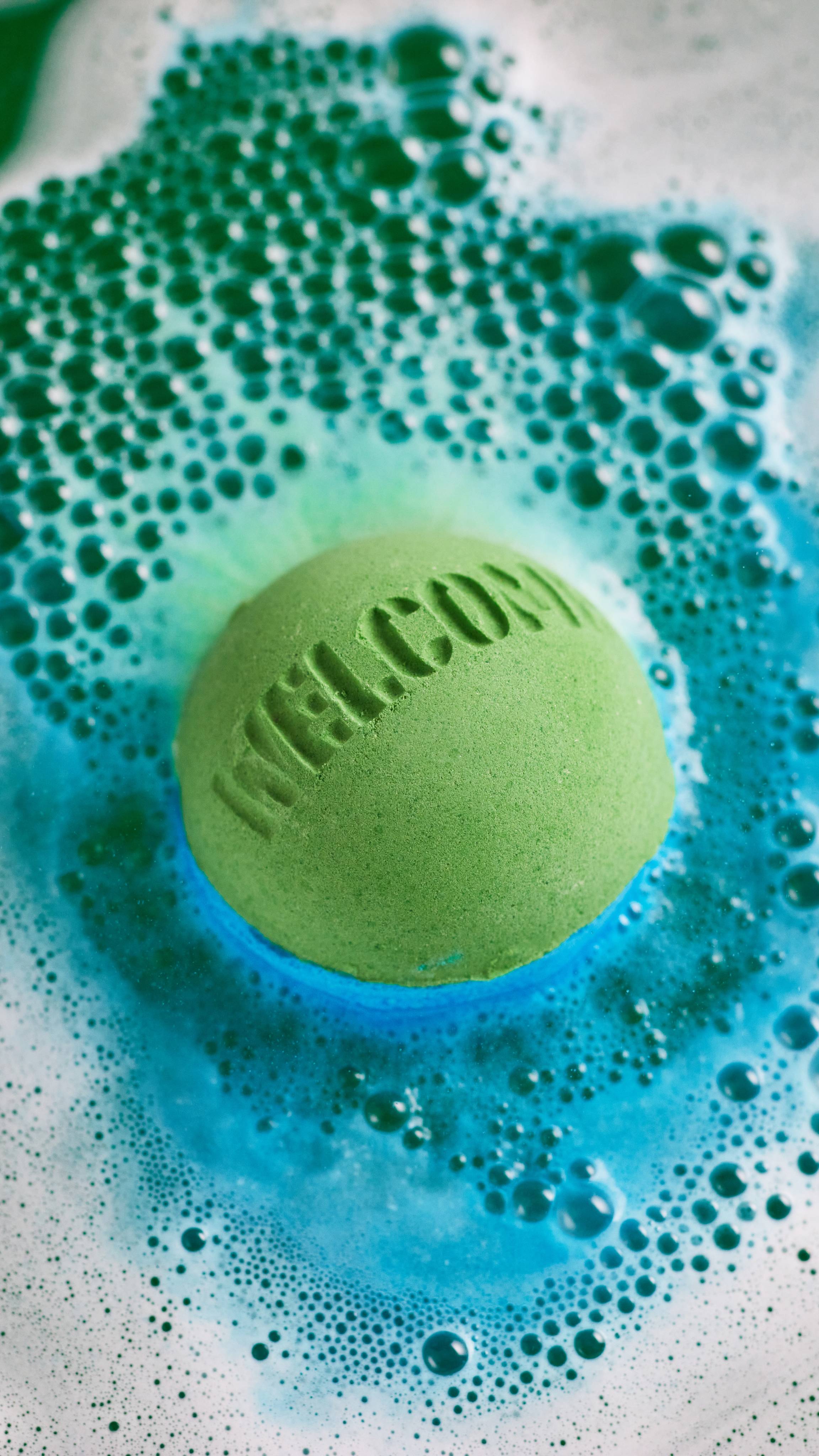 Welcome bath bomb floats among a carpet of bubbles in colourful deep-sea green, blue and teal swirls.