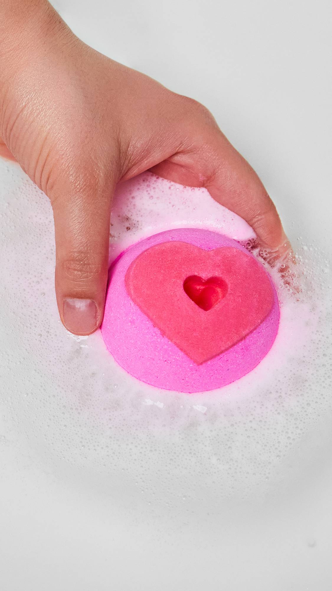 The image shows a close-up of the model holding the Whispering Heart bath bomb just on the surface of the water. 