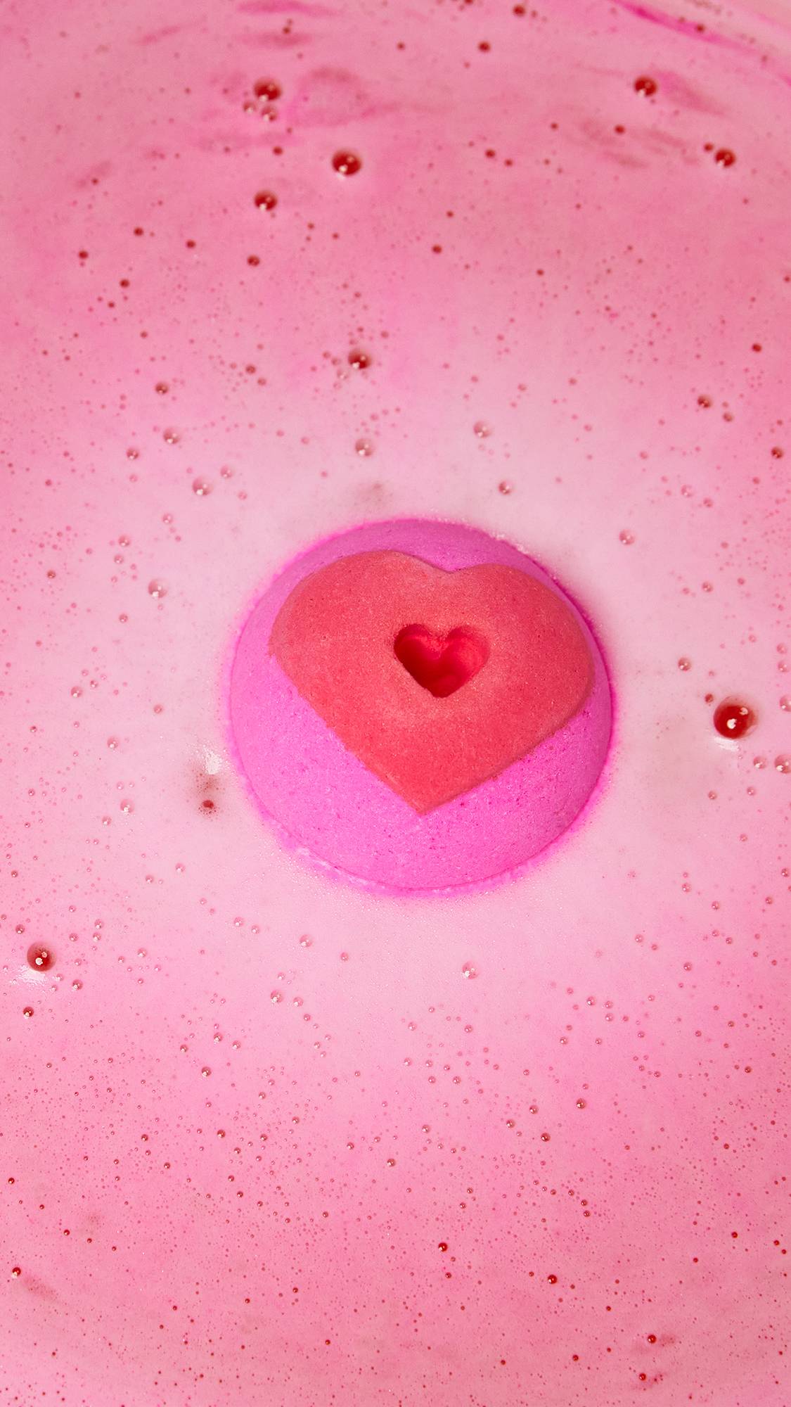 The Whispering Heart bath bomb sits on the water with the heart and hole on top as it is surrounded by thick, pink, foamy bubbles.