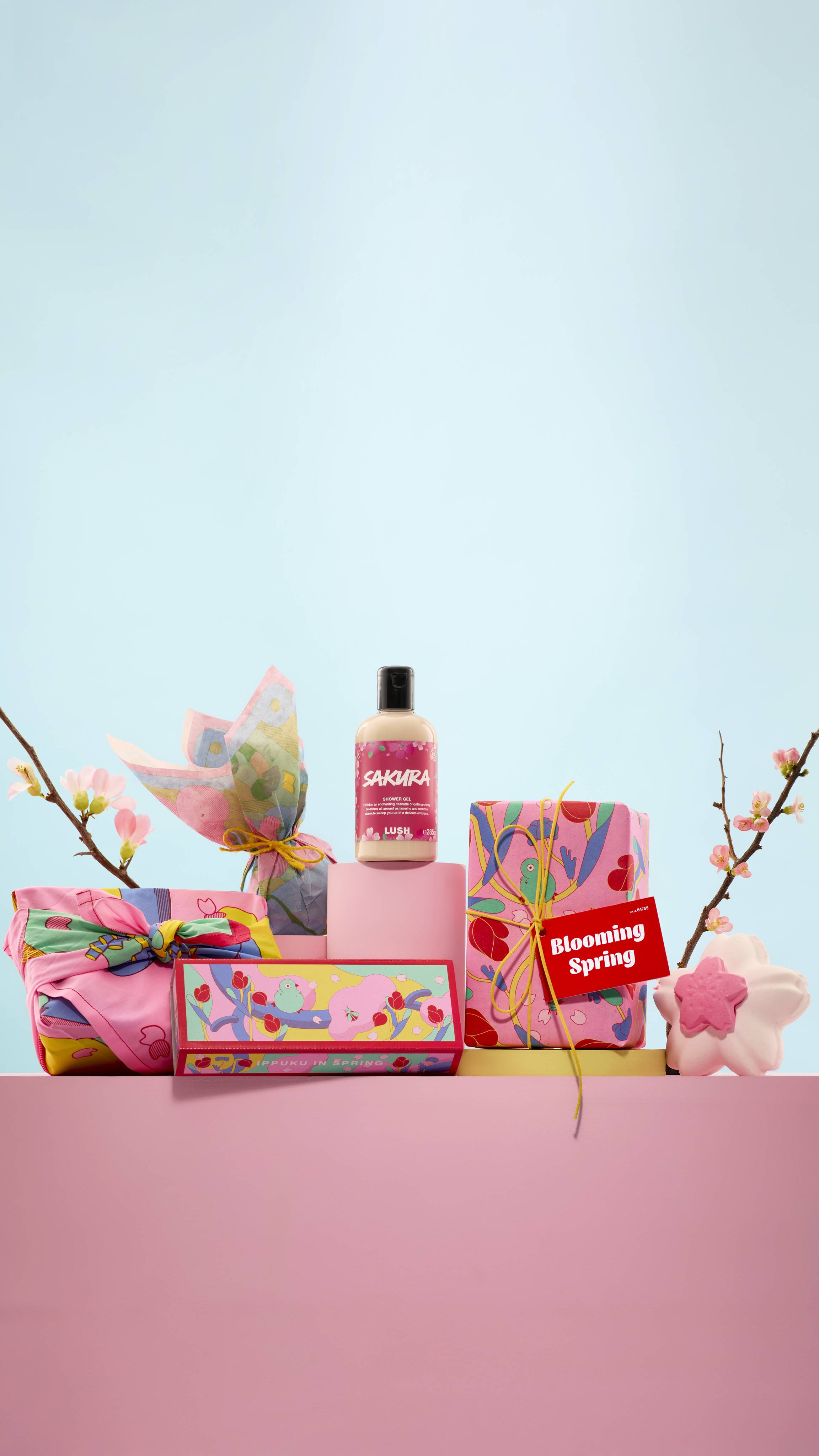 The image shows the whole LUSH "White Day/Sakura" range on pink podiums. It features both Sakura-themed gifts, a knot wrap, Lokta paper, a bath bomb and a bottle of shower gel, with Cherry Blossom branches.