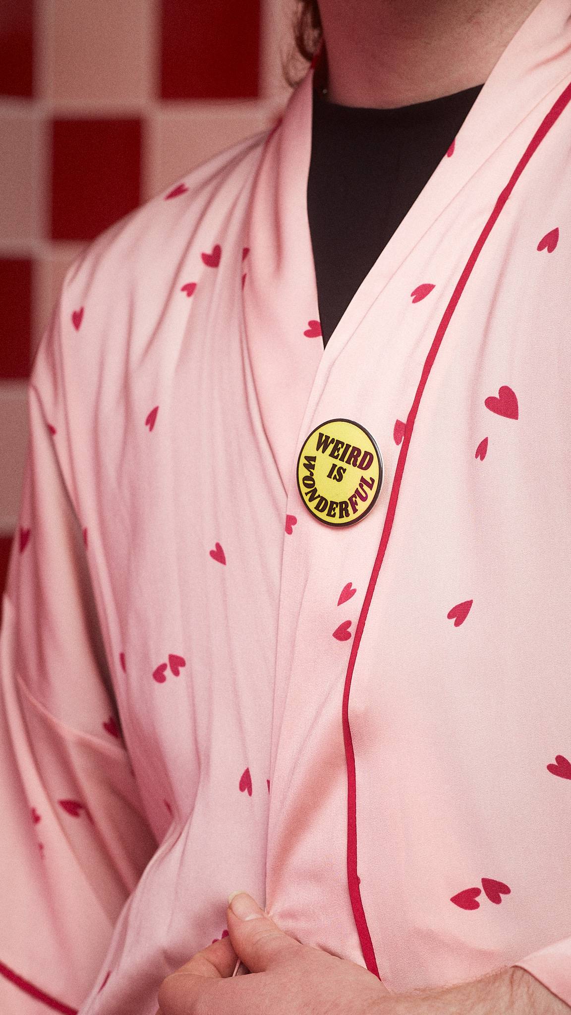 Model is wearing a white robe dotted with hearts. A bright yellow badge sits on the lapel with the words "Weird is Wonderful" in black.