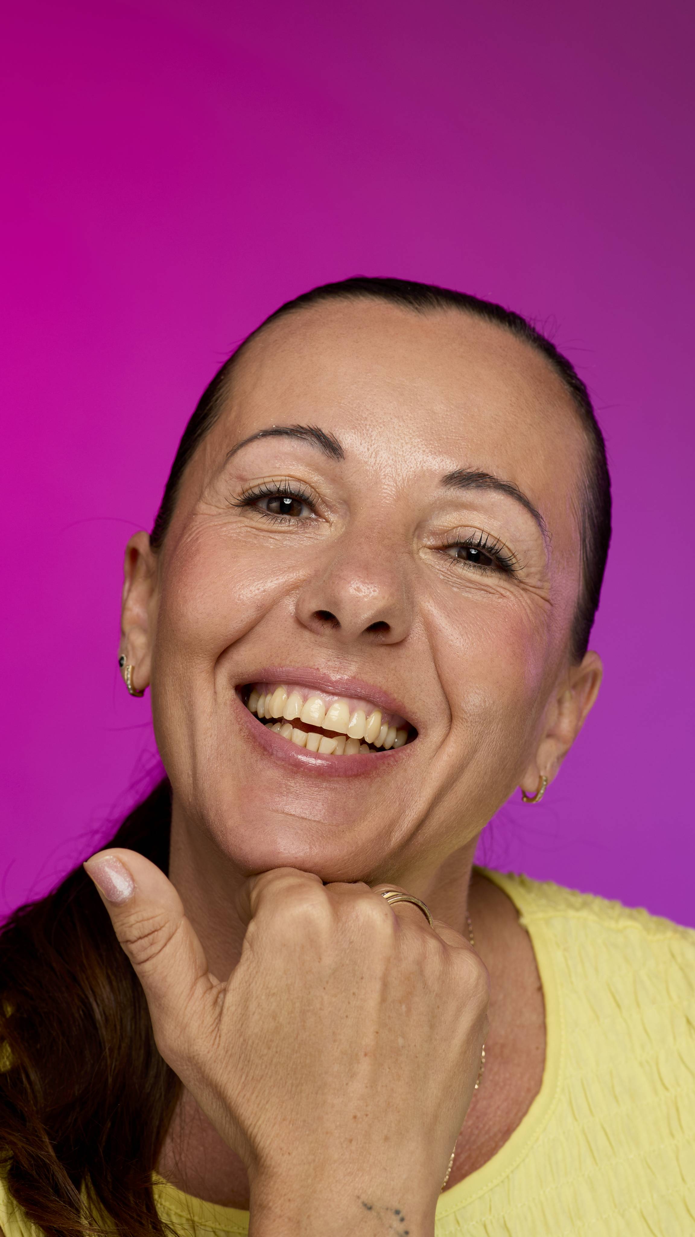 A smiling model with fair skin and tied-back dark hair is shown from the neck up, with their chin resting on their hand, on a vibrant pink background. They smile, showing off the rich, mauve-brown Willow lipstick with a subtle shimmer. 