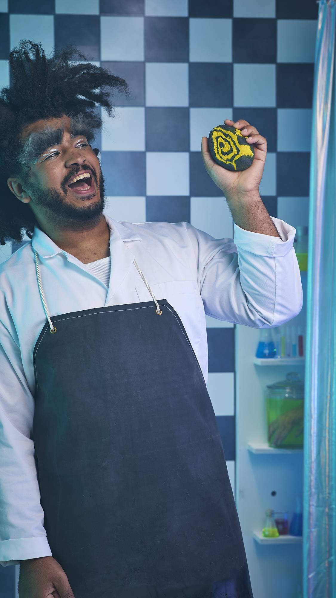The image shows the model dressed as a mad scientist complete with a black apron and wild hair. They are standing on a black and white checkered background and are holding the Witchy Woo bubble bar in their hand.